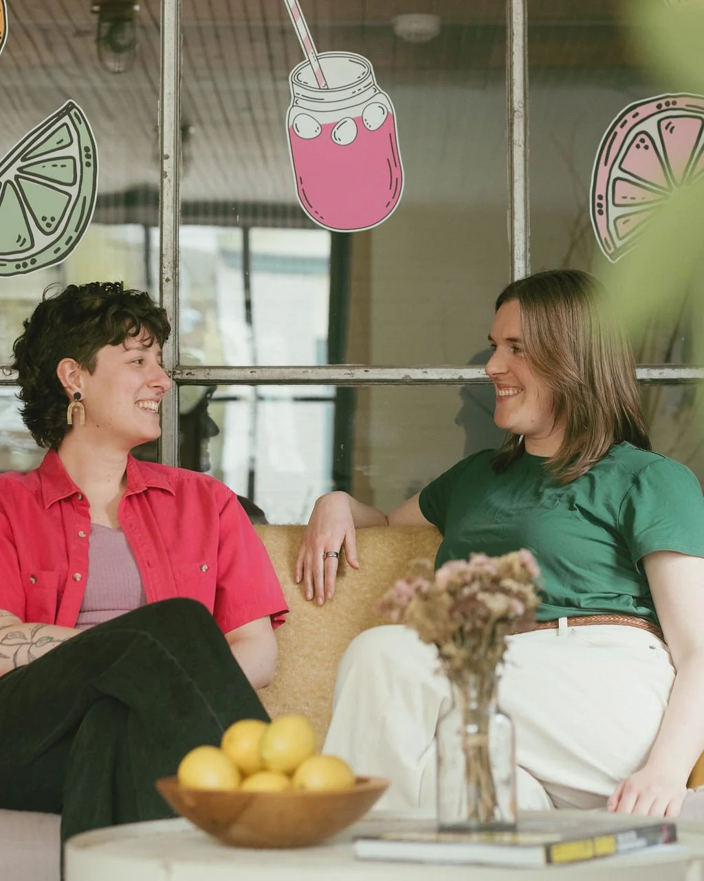 Two women sitting on a yellow couch smiling and talking in the Lemonade Co. office with lemon and drink illustrations on the window behind them.