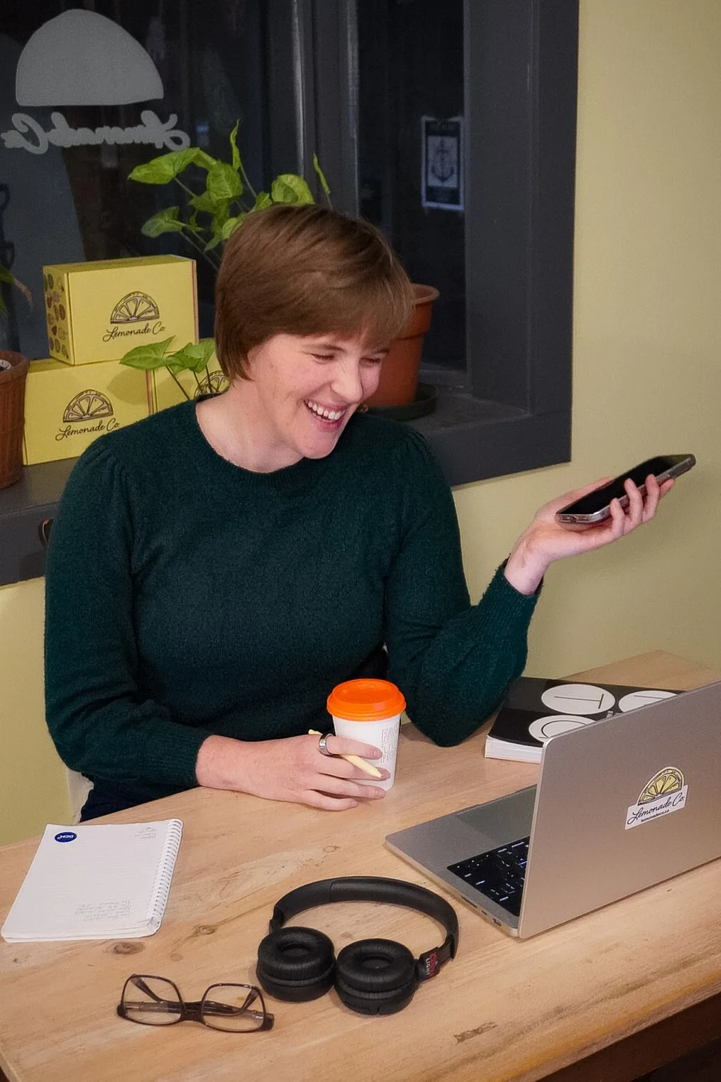 A woman with short reddish hair is sitting at a wooden table, smiling while looking at her phone. She has a notebook, headphones, a pair of glasses, a laptop with a Lemonade Co sticker, a coffee cup, and a pen on the table. Behind her, there are lemon-themed boxes and pots with green plants on a windowsill.