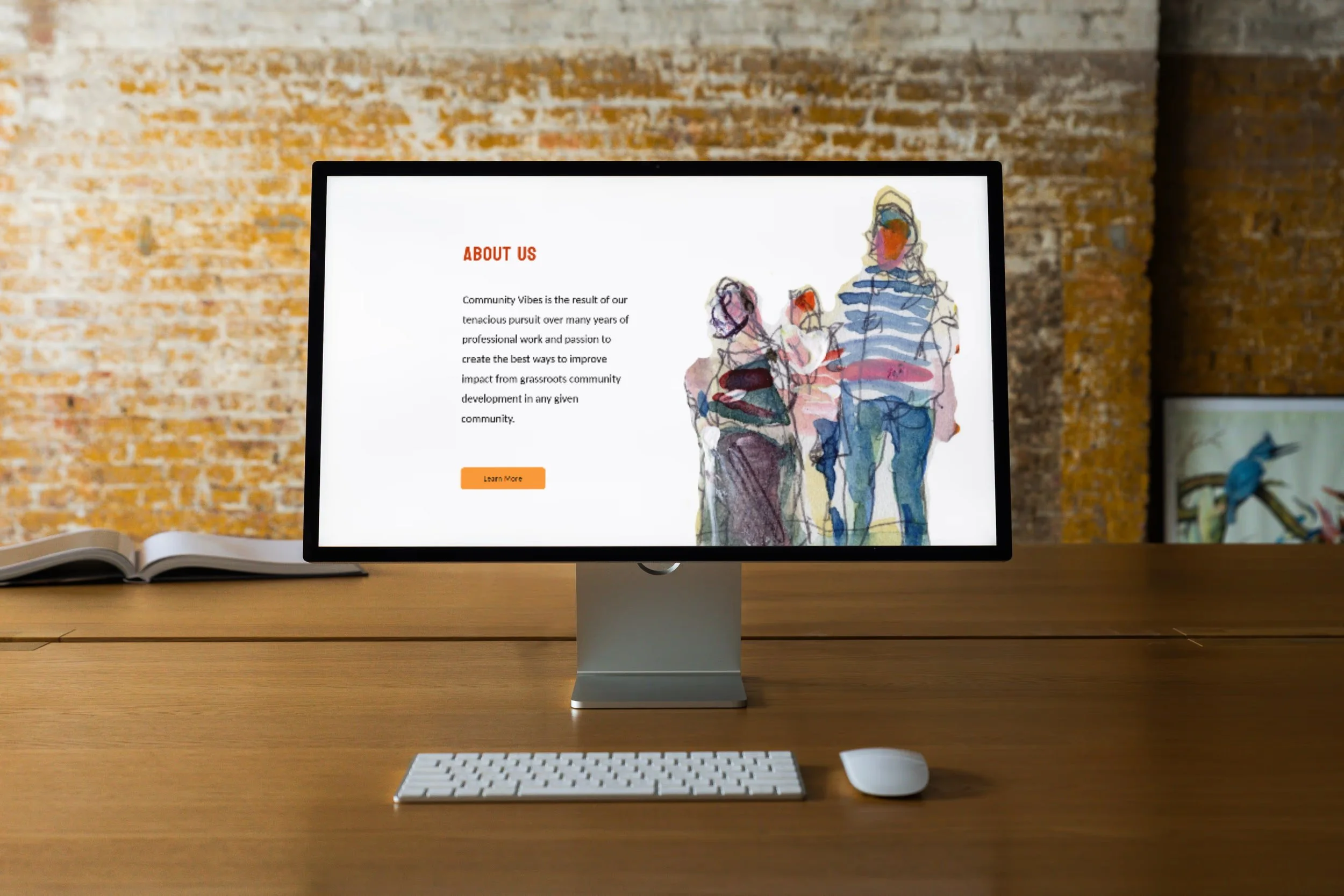 Computer monitor on wooden desk displaying a webpage with an illustration of people and text titled About Us. The screen is showing a mock-up of a website design by Lemonade Co.