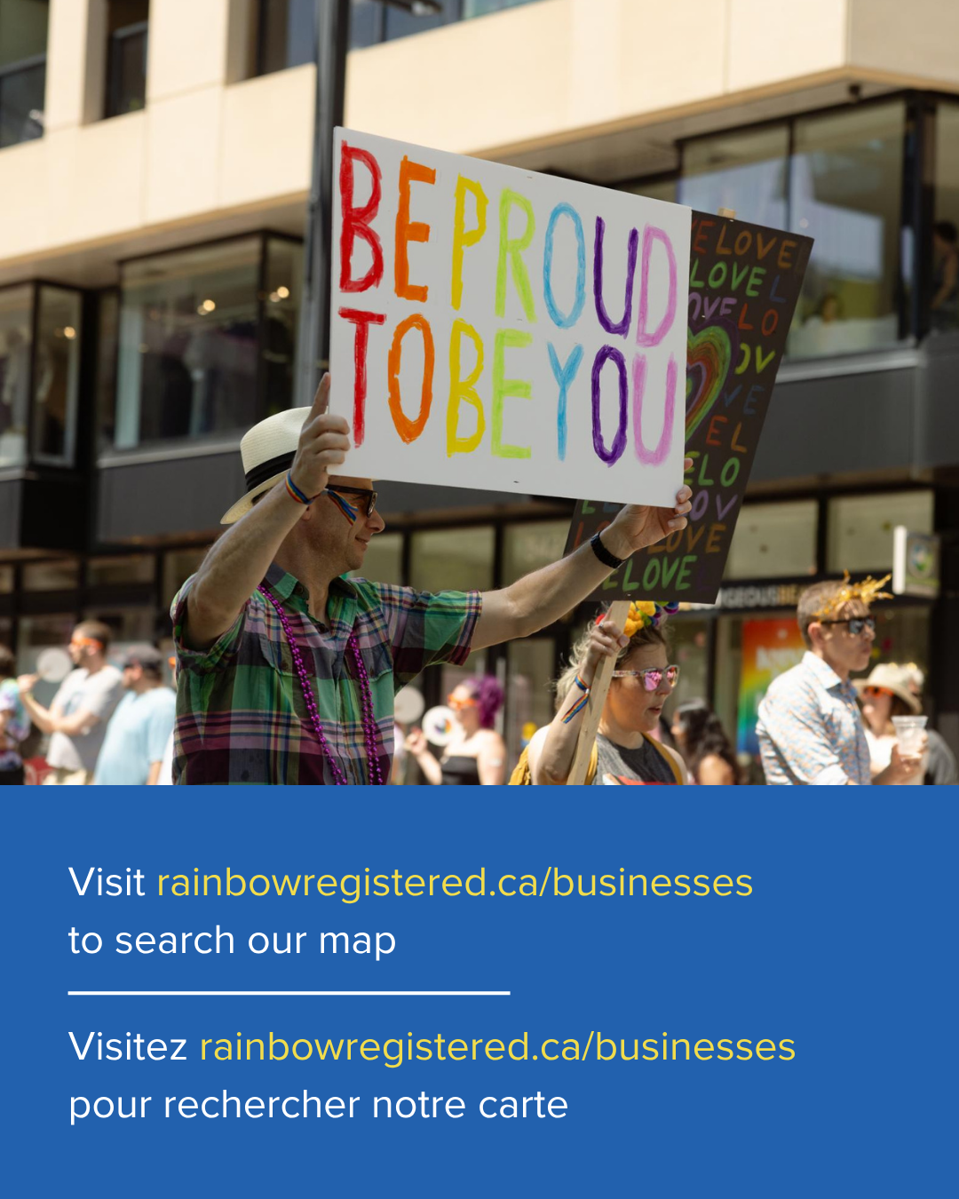 A person holding a rainbow-colored sign that says 'Be Proud To Be You' during a pride parade, with other parade participants in the background.