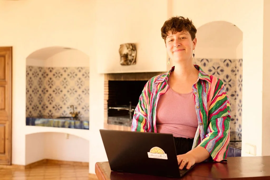 A woman with short, curly brown hair sitting at a table with a laptop, smiling in a warmly lit room with a fireplace and decorative wall niches.