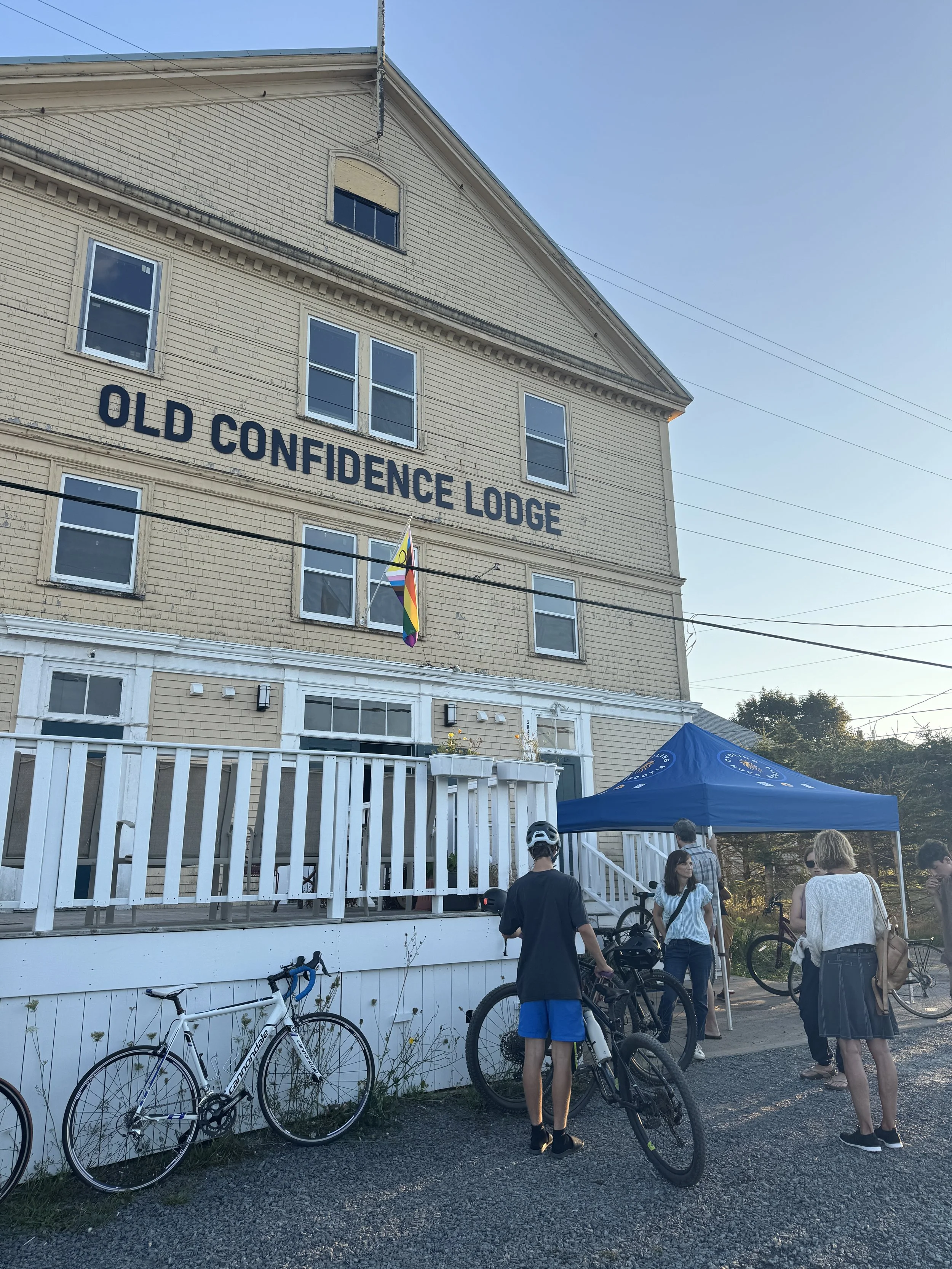 People gather outside a beige, multi-story building called 'Old Confidence Lodge,' with bicycles parked against the front fence and a blue canopy tent nearby, during daytime.