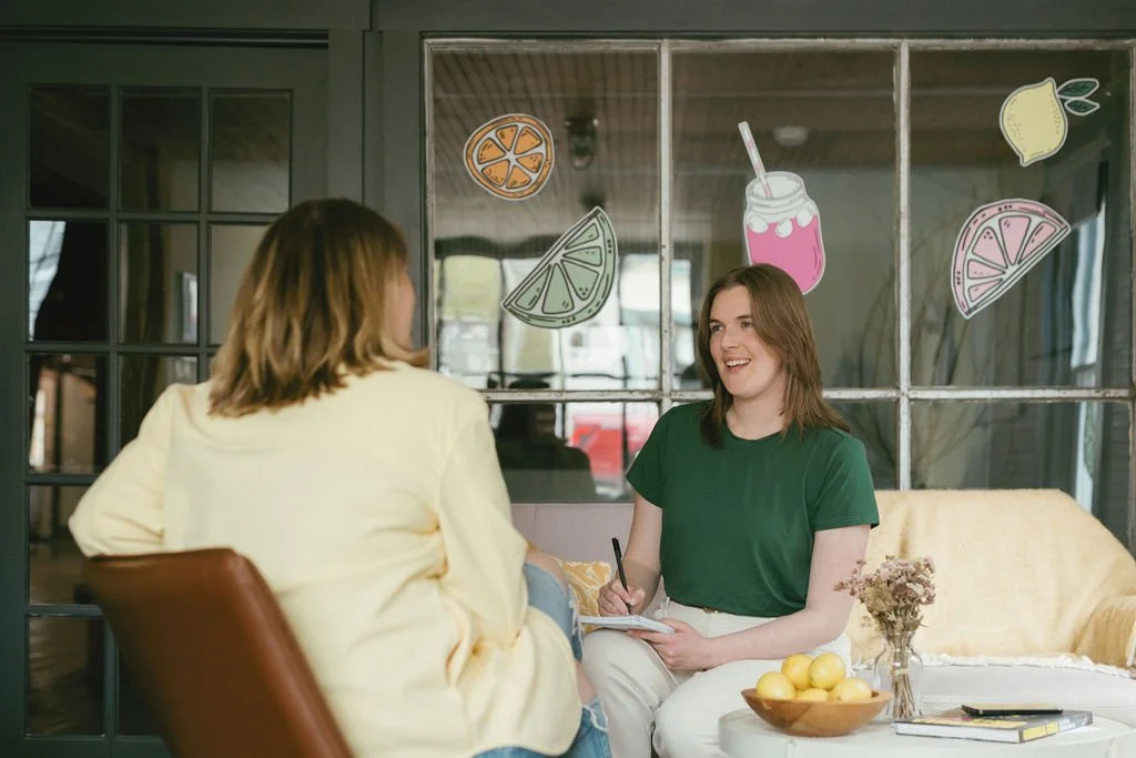 Emily, a person with brown hair and a green shirt on, is talking to a client sitting across from her