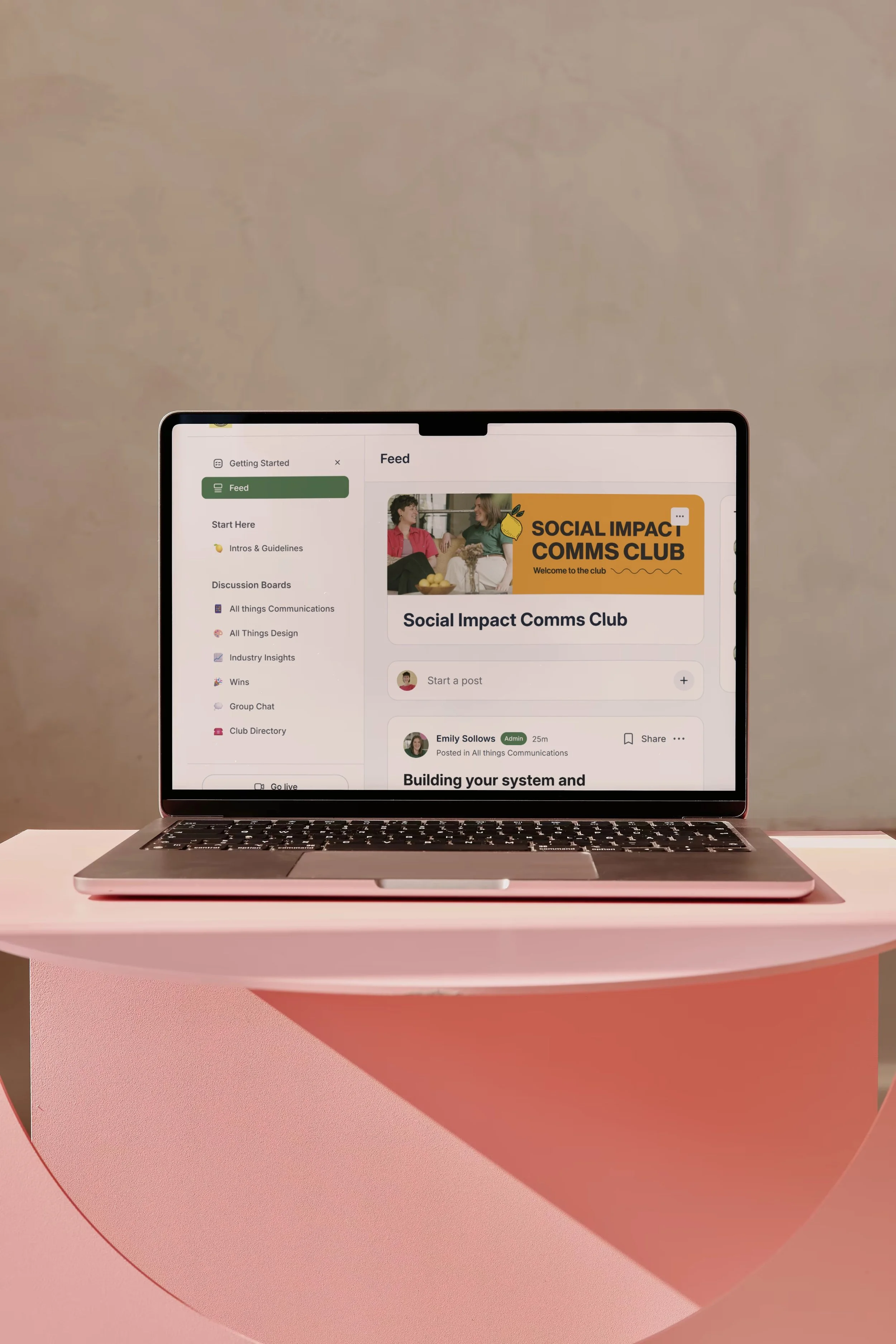 A laptop on a pink circular table displaying a social media or communication app with a feed, discussion boards, and an event for a Social Impact Communications Club.