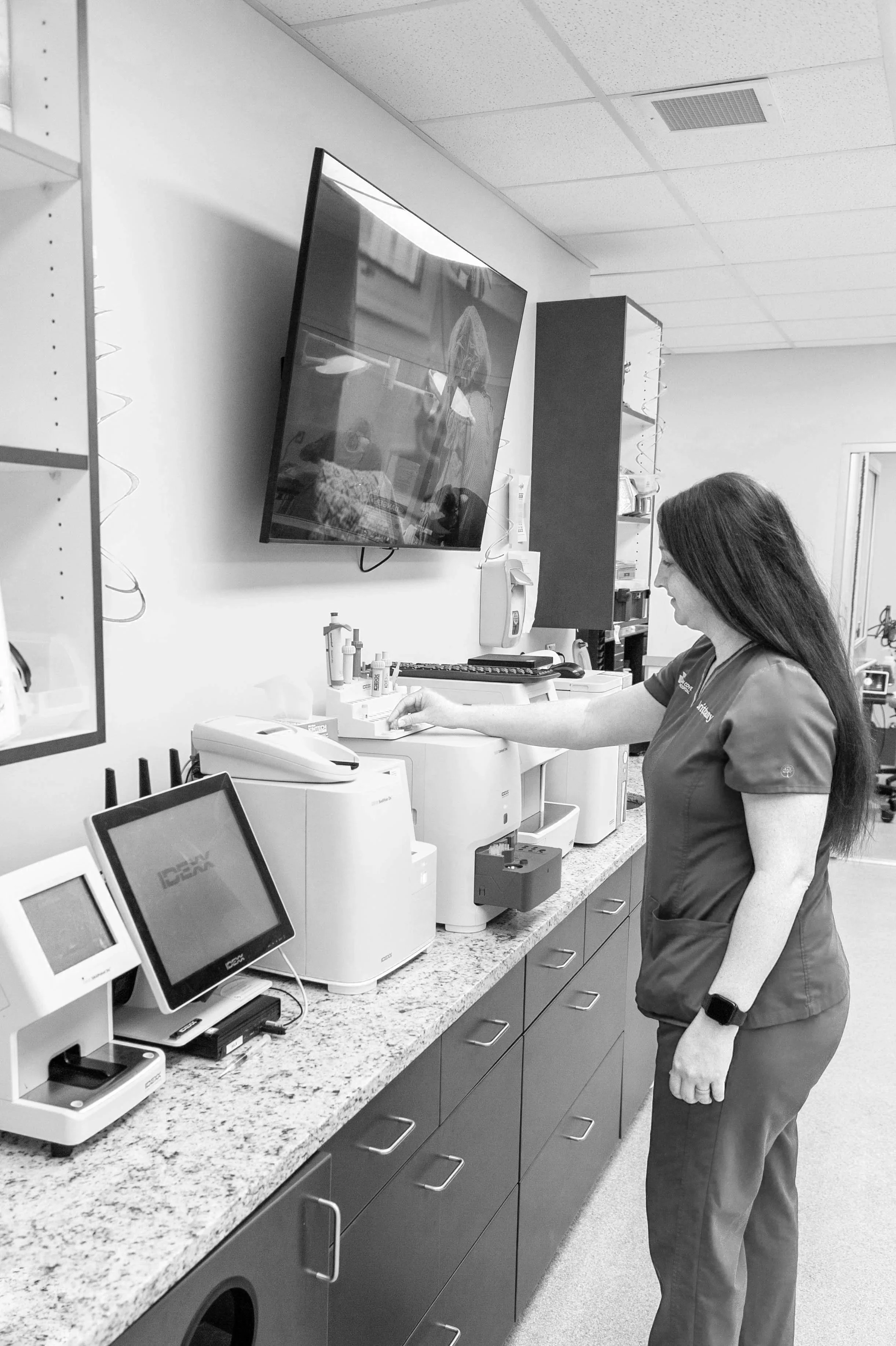 A female healthcare worker in scrubs standing at a medical supply counter in a clinic, reaching for items on the counter, with medical equipment and a wall-mounted television in the background.