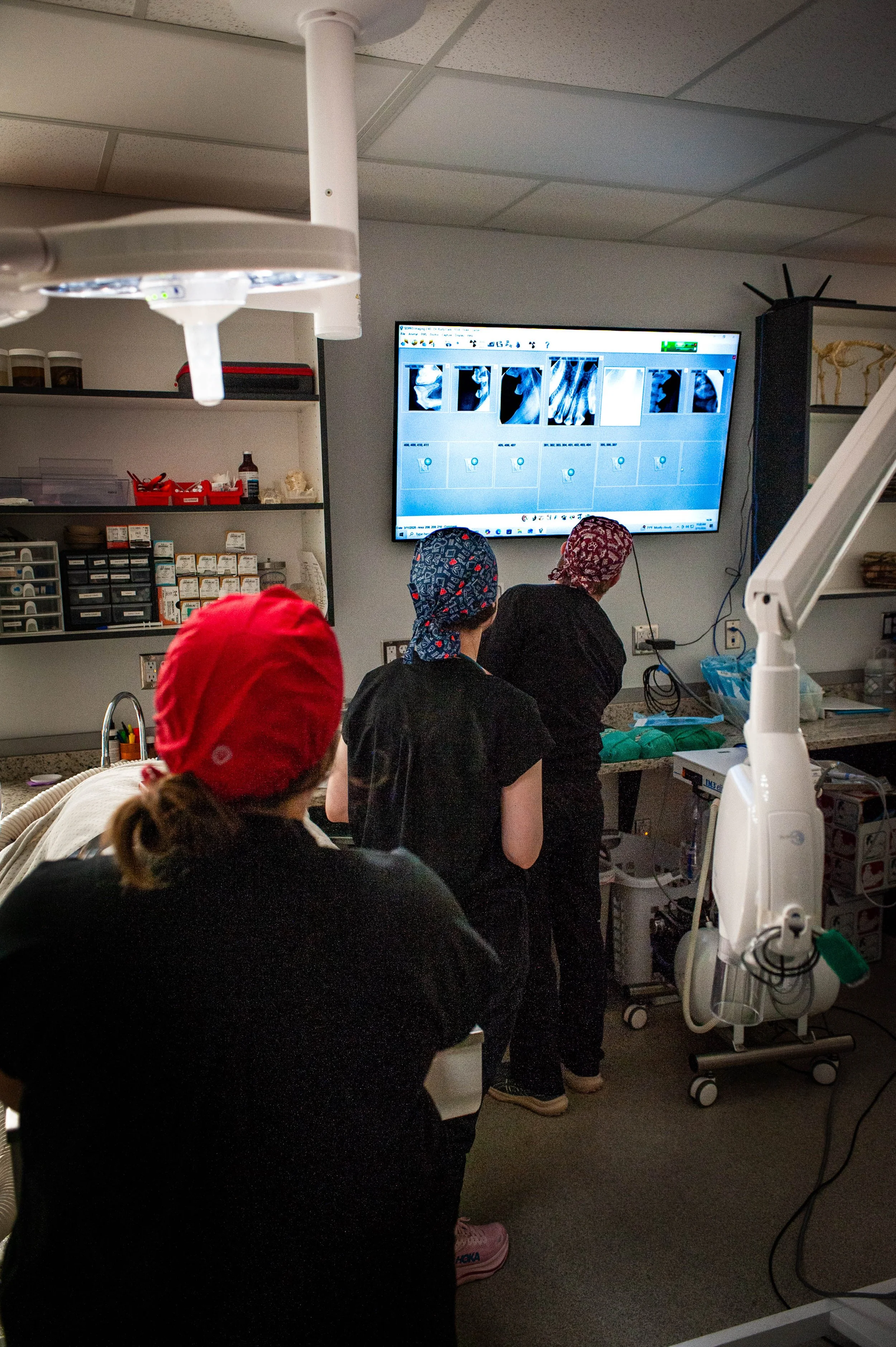 Medical professionals in scrubs and surgical caps review X-ray images on a large monitor in a hospital or clinic setting.