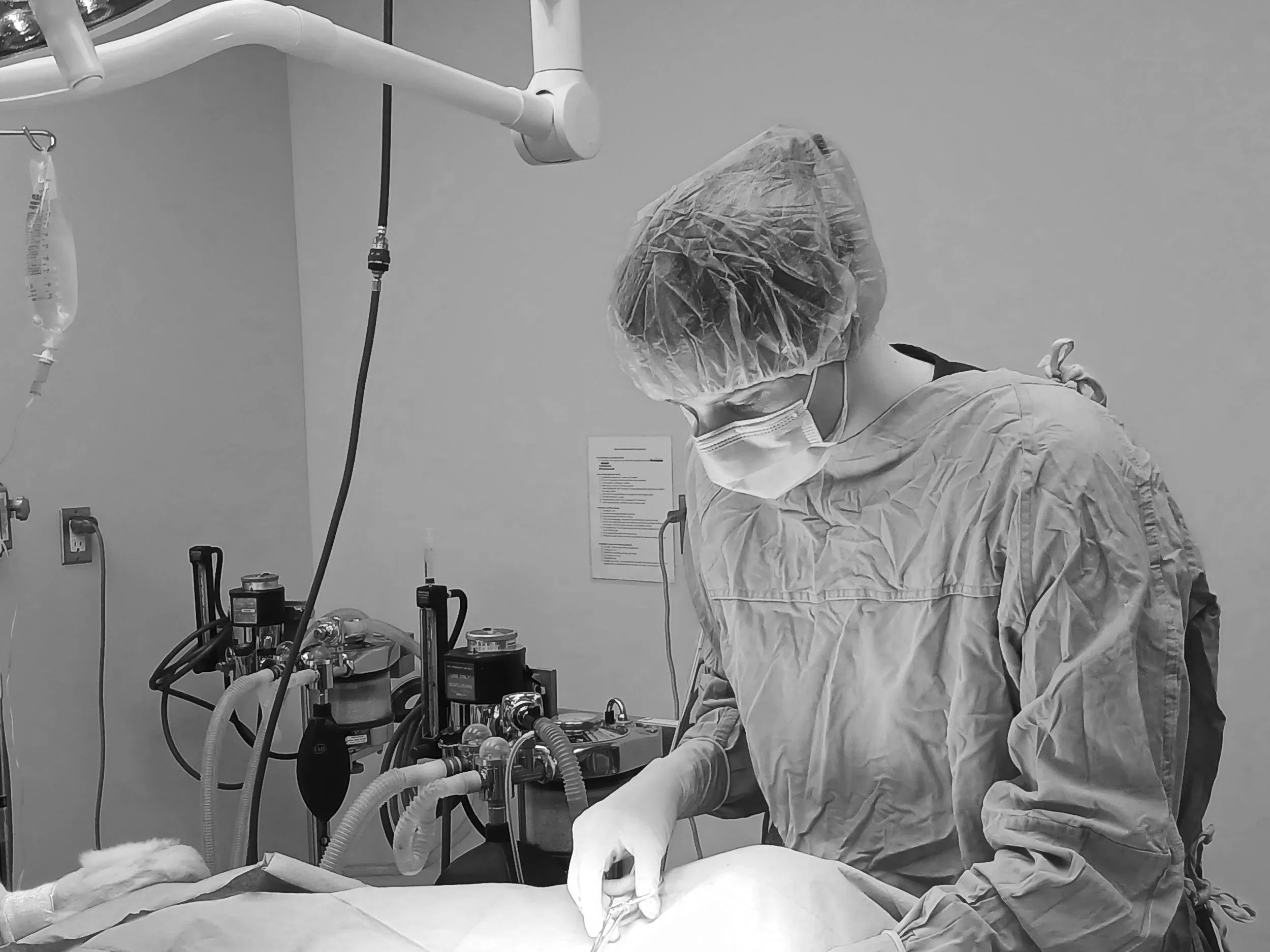 A medical professional dressed in scrubs, mask, hair cover, and gloves performing a procedure in an operating room.