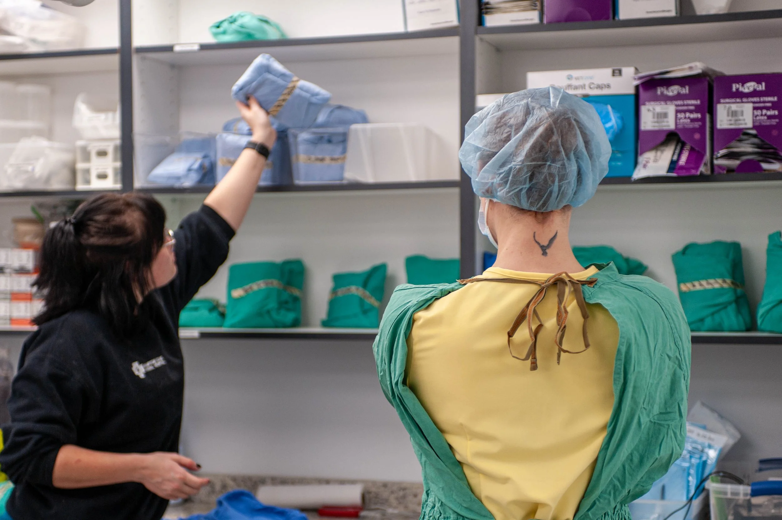 Two healthcare workers in protective gear, one woman with glasses and dark hair and another person with a face mask and blue hair cover, working in a storage room with medical supplies.