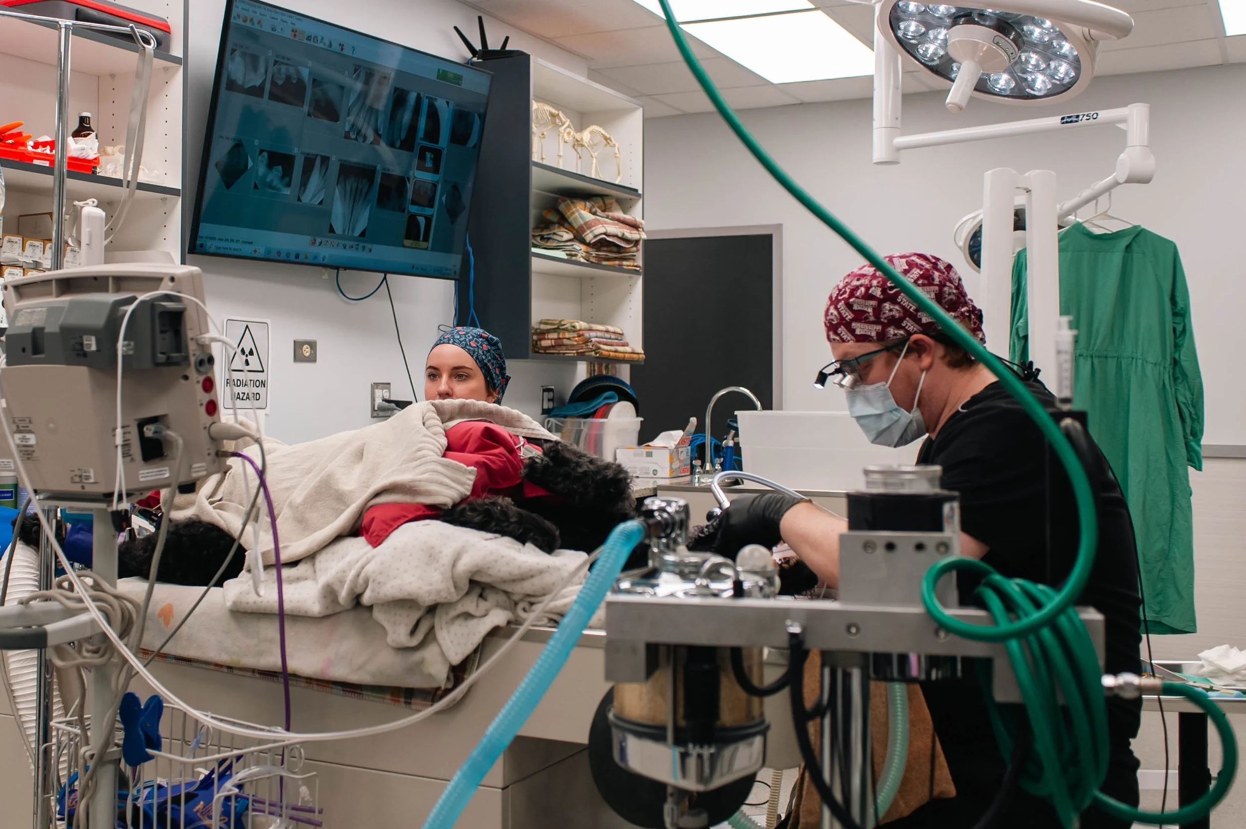 A woman lying in a hospital bed with a blanket, receiving medical care from a healthcare professional in an operating room. The healthcare worker is wearing a mask, gloves, and a surgical cap, working with medical equipment. There are surgical lights