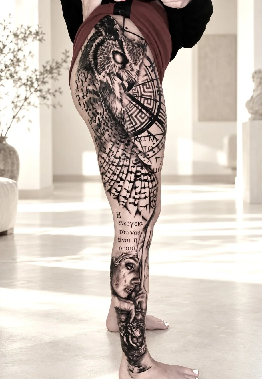 Close-up of a person's leg with detailed black and gray tattoos, including an owl, a face of a woman, Greek text, and various symbols, in a brightly lit room.