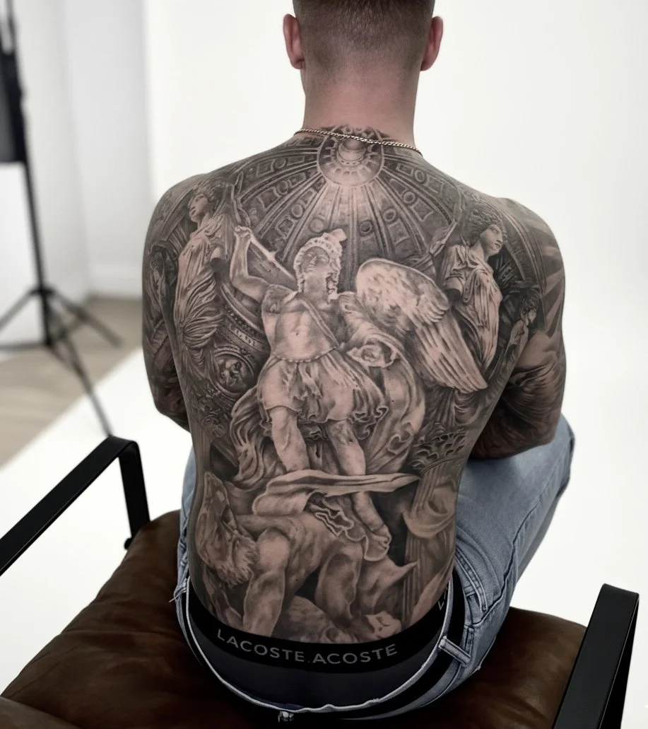 A man with intricate black and gray tattoos covering his back, depicting classical-style sculptures and figures, sitting on a brown cushion.