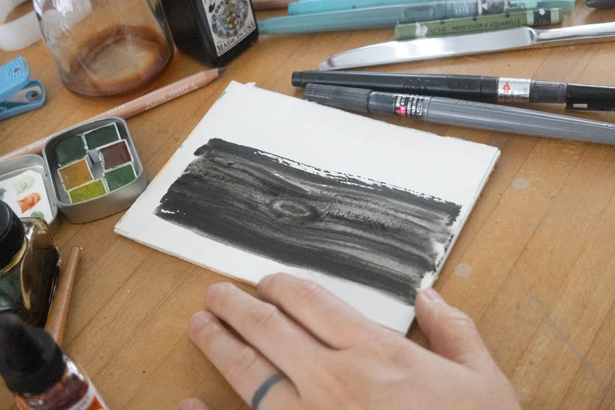 Open sketchbook with small paint and texture studies on a cluttered desk covered in pens and brushes.