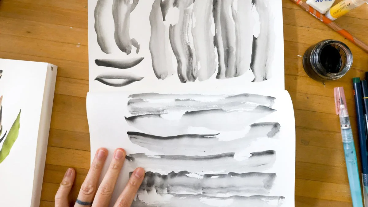 Hand holding open a sketchbook filled with loose black ink wash strokes and texture studies on a wooden desk.