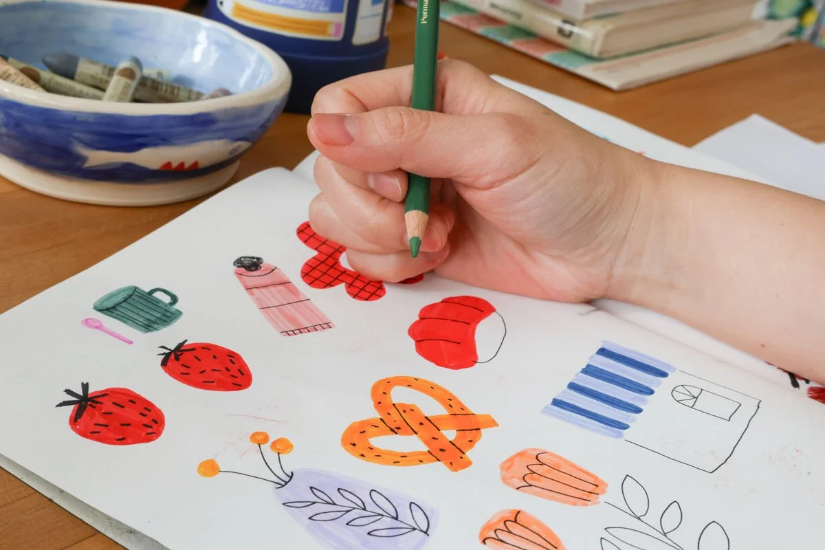 Hand drawing simple colorful icons—strawberries, pretzel, cup and flowers—in a sketchbook.