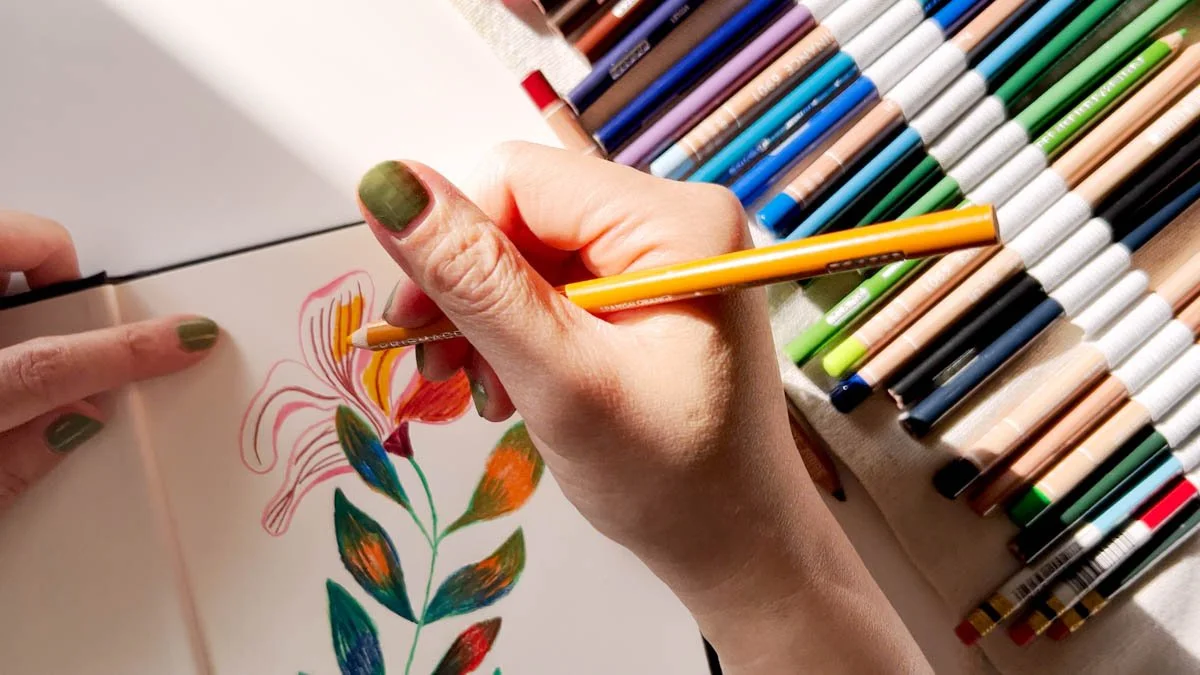 Hand coloring a bright flower in a sketchbook beside a neatly arranged roll of colored pencils in sunlight.