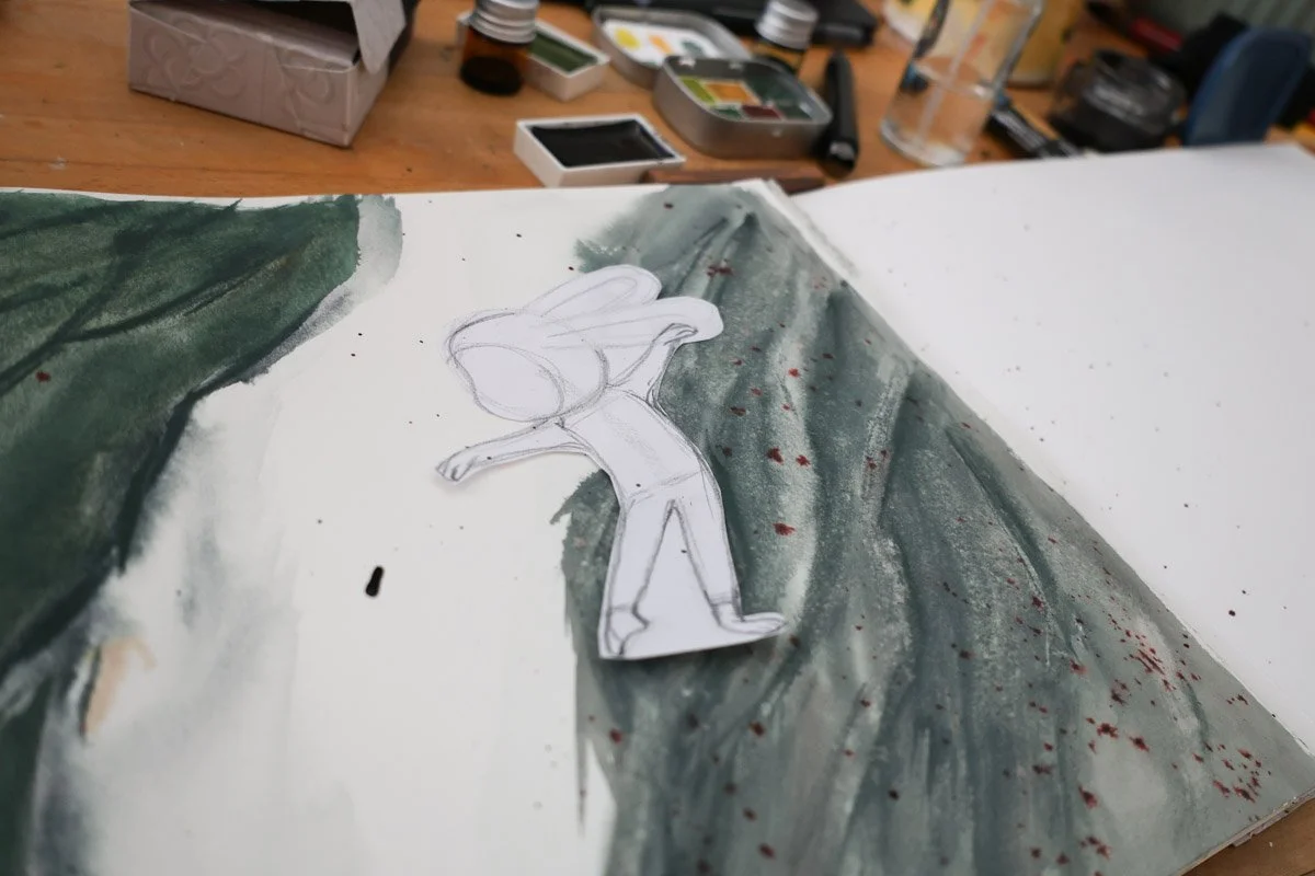 Paper cut-out bunny character placed on top of a loose green watercolor background in a sketchbook.
