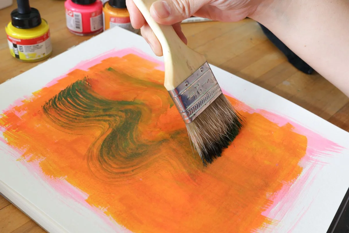 Hand using a wide paintbrush to pull green, wavy strokes through bright orange and pink paint on paper.
