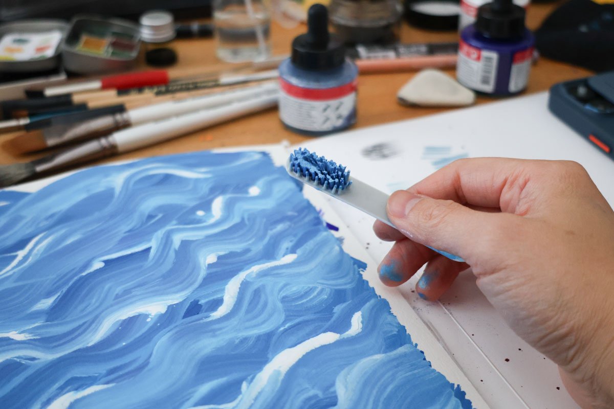 Hand holding a paint-covered toothbrush over a blue wavy painted surface, with inks and brushes nearby.
