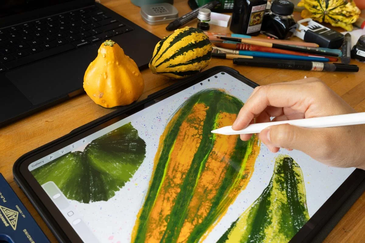 Hand drawing a striped gourd on an iPad with a stylus, surrounded by real gourds and art supplies.