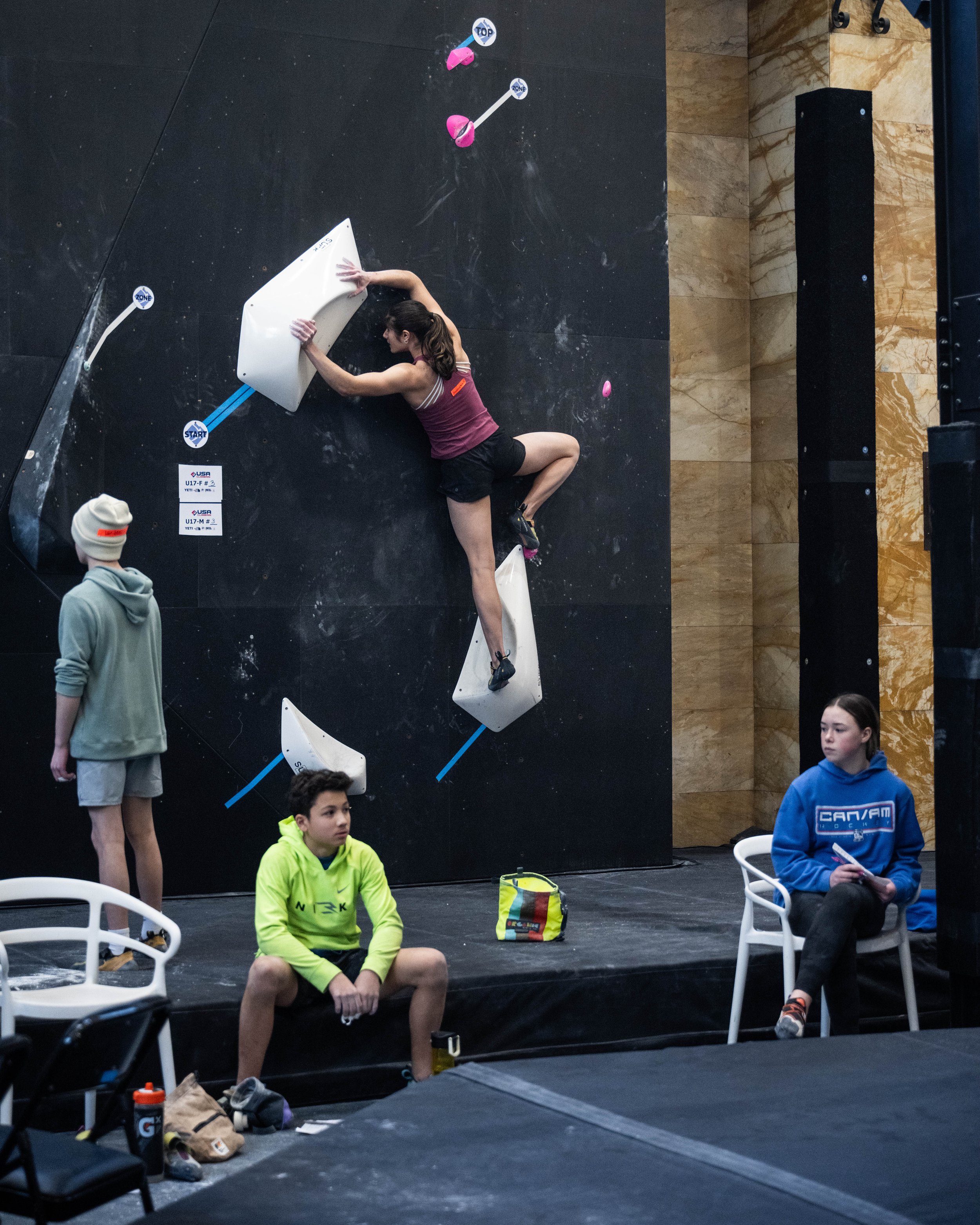 Indoor rock climber practicing bouldering on a black climbing wall with white and pink holds; two seated spectators and one standing observer.