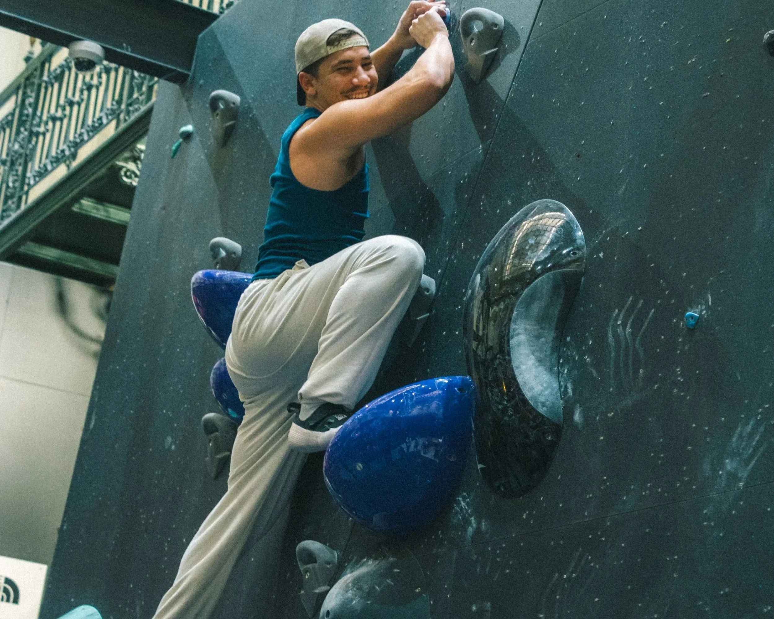A young man smiling while climbing an indoor rock wall, wearing a beige cap backwards, a blue tank top, and khaki pants. Indoor rock climbing.