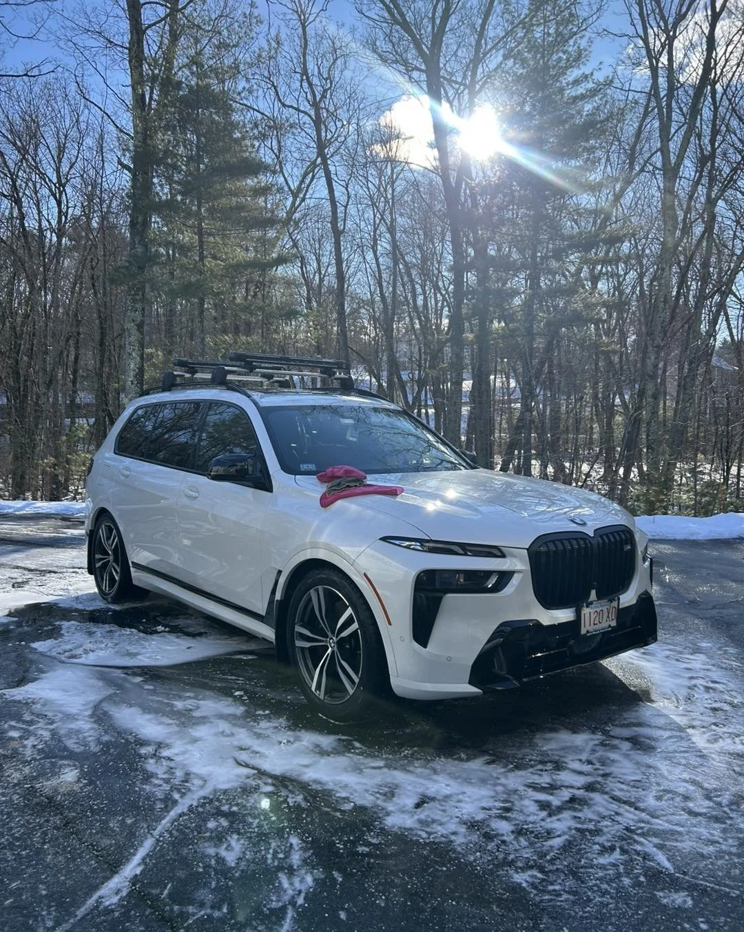 A white SUV parked on icy ground in a snowy wooded area with leafless trees, sunlight shining through the branches, and a pink item on the car's hood.