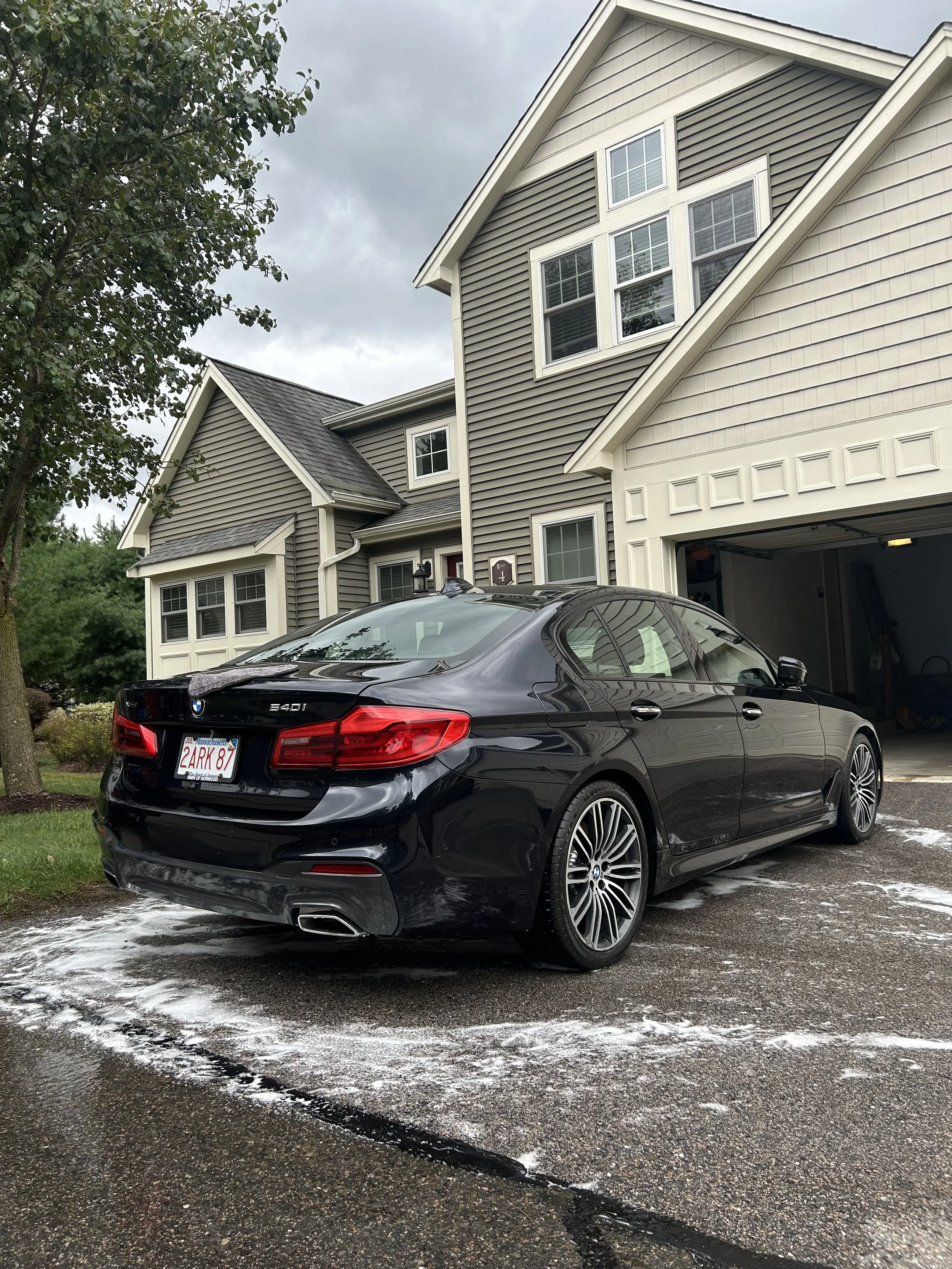 A black BMW 540i parked in a driveway next to a beige and gray house with a garage. The driveway has soap or foam residue from washing the car, and the sky is overcast.