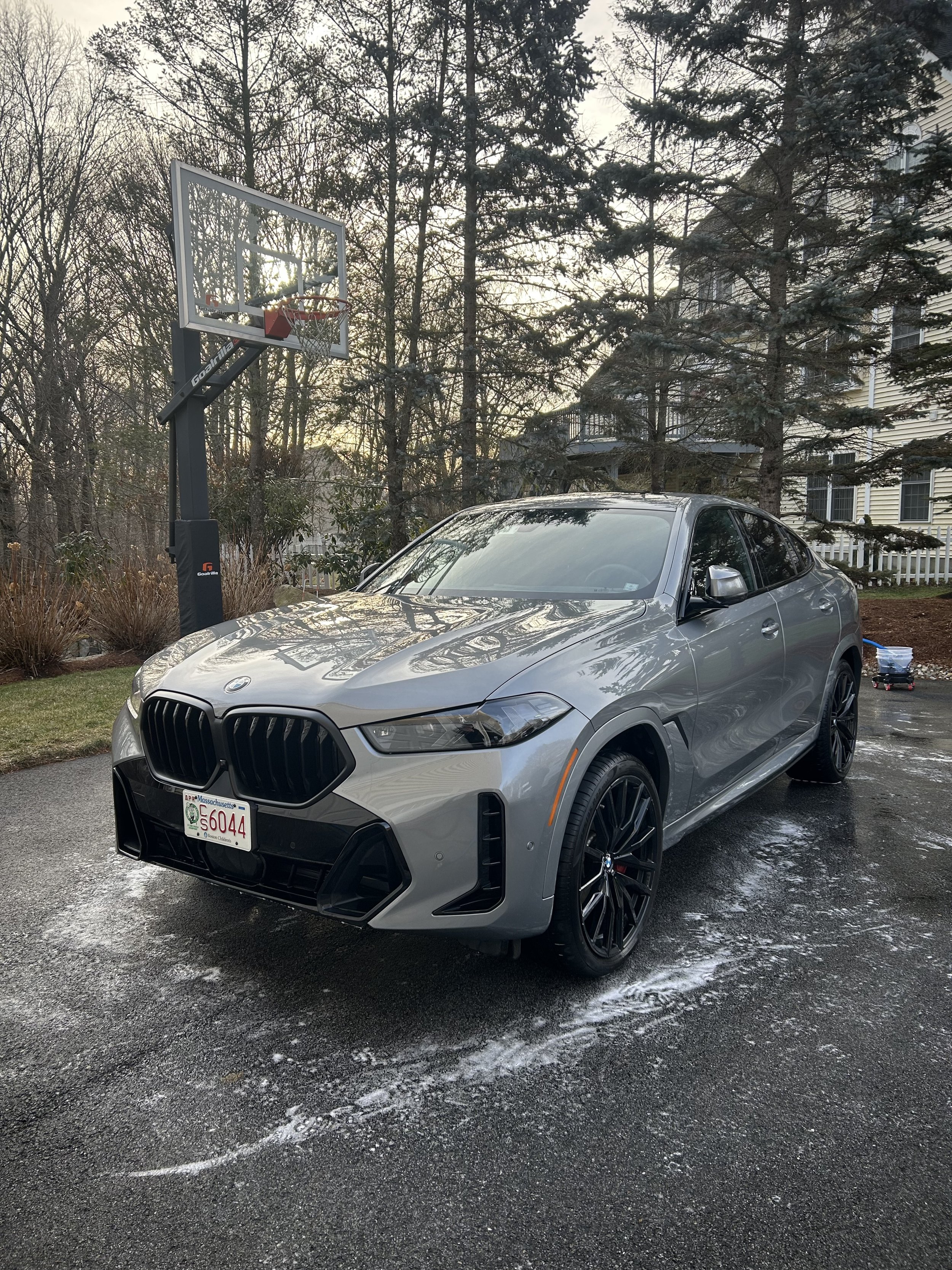 A silver BMW SUV parked on a driveway next to a basketball hoop, with a house and trees in the background.