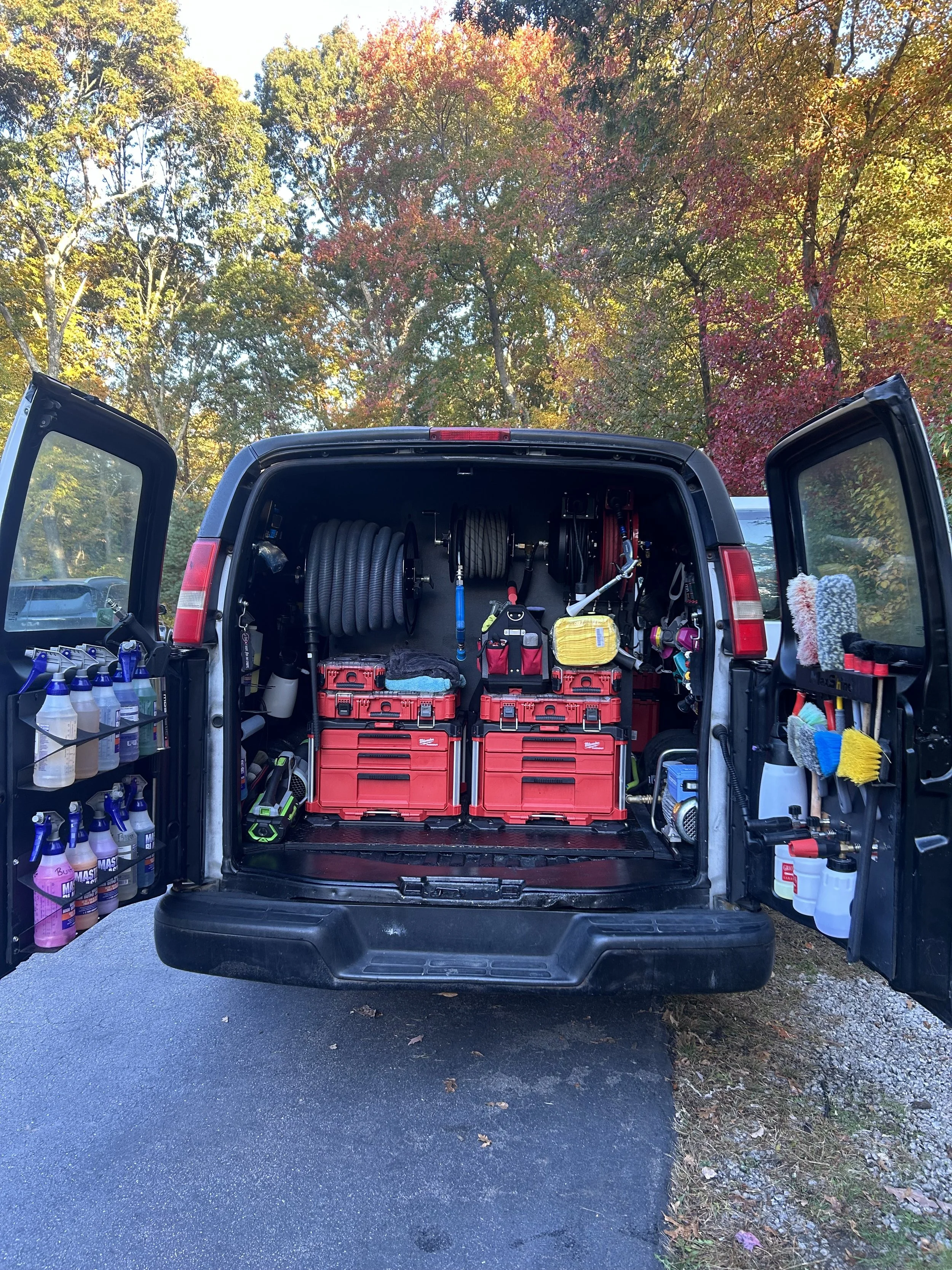Open van trunk showing organized cleaning and maintenance equipment including spray bottles, hoses, toolboxes, microfiber cloths, brushes, and cleaning tools in a natural outdoor setting with colorful autumn trees in the background.