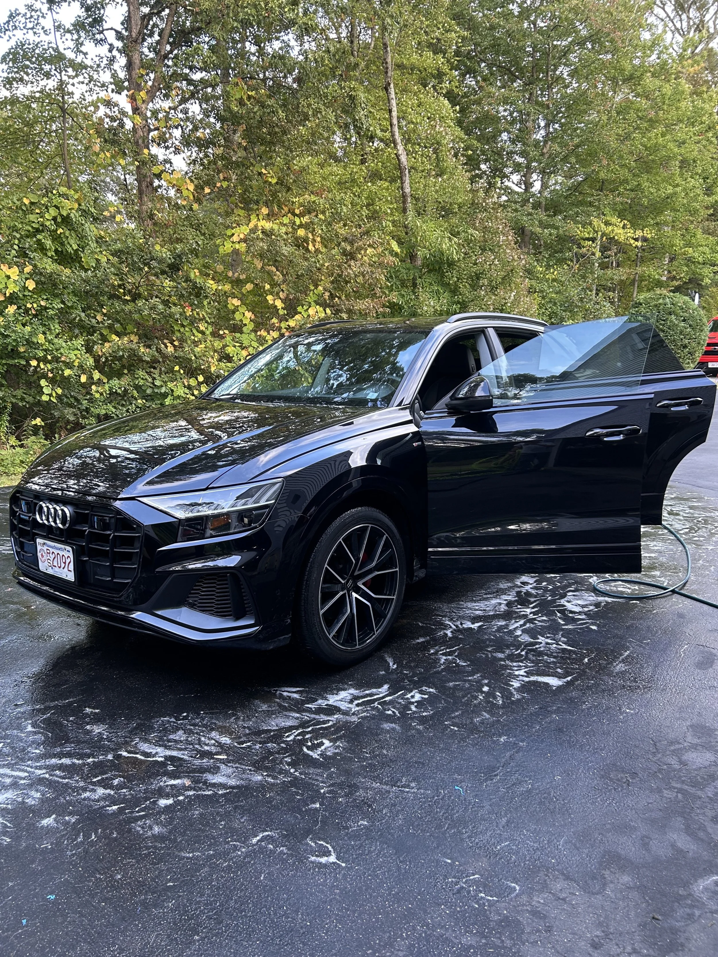 A black Audi sedan parked on a wet driveway with soap and water, with its driver side door open and an outdoor wooded area in the background.