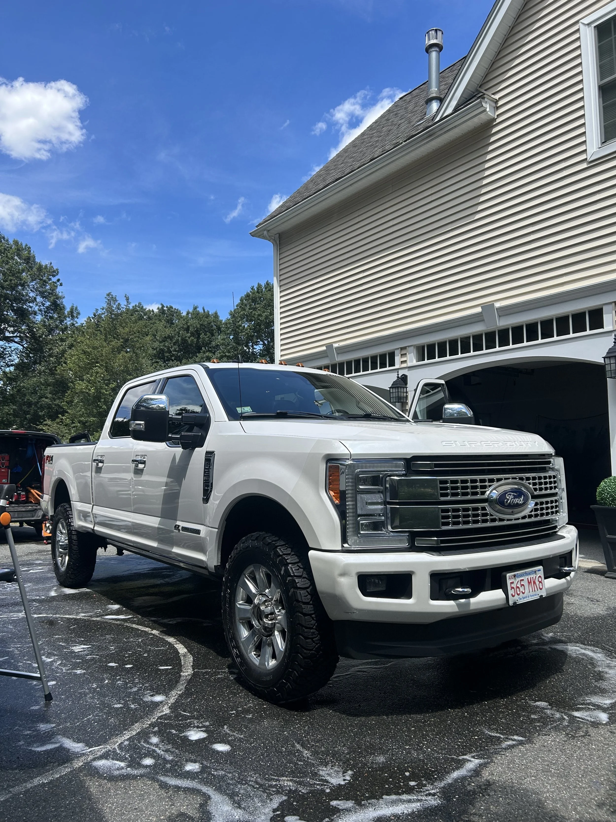 White Ford pickup truck parked on a driveway with soap and water, next to a house with white siding and garage, under a blue sky with scattered clouds.