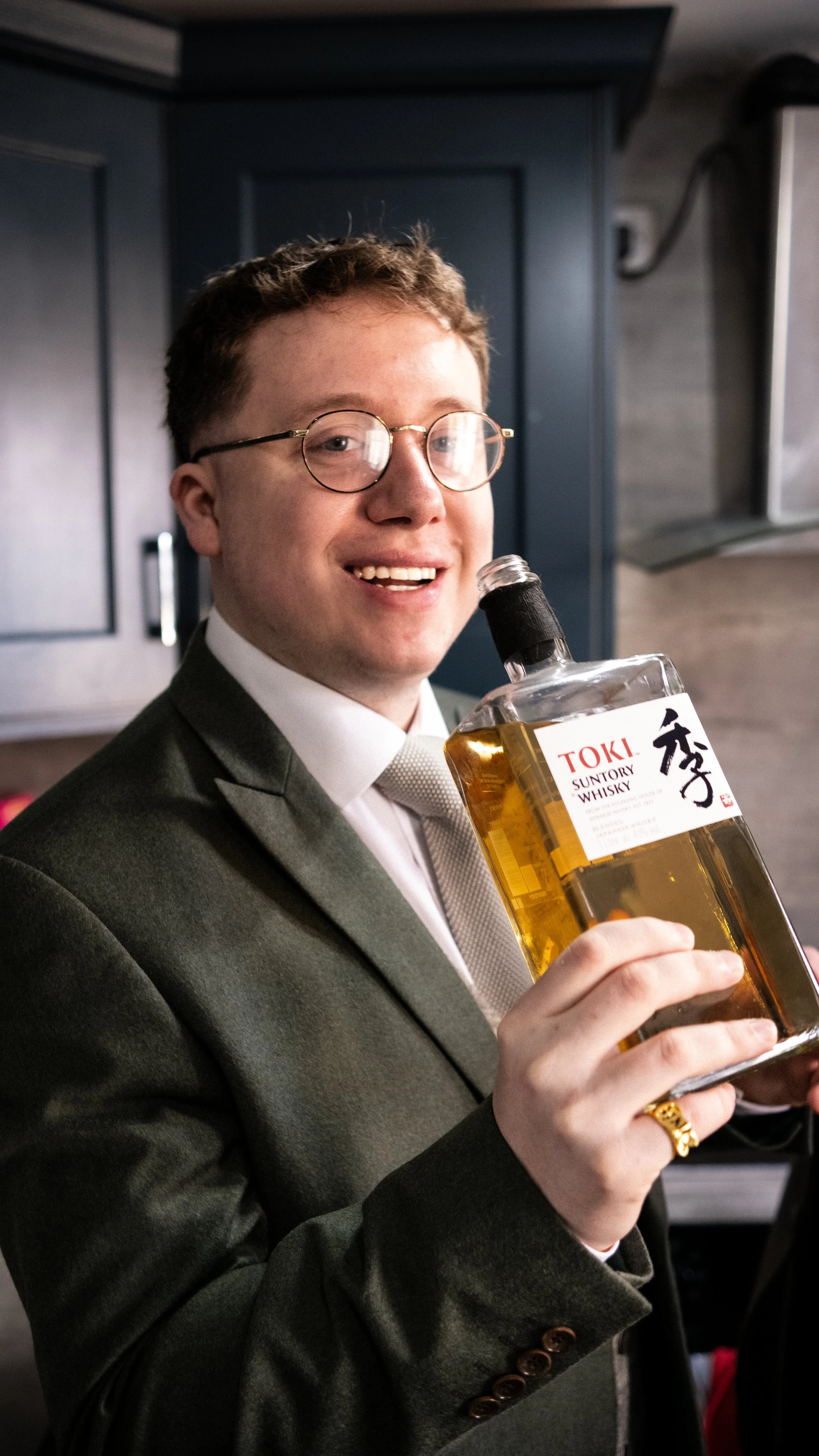 A young man in a suit and tie holding a bottle of Toki Suntory Whisky, smiling at the camera in a modern kitchen.