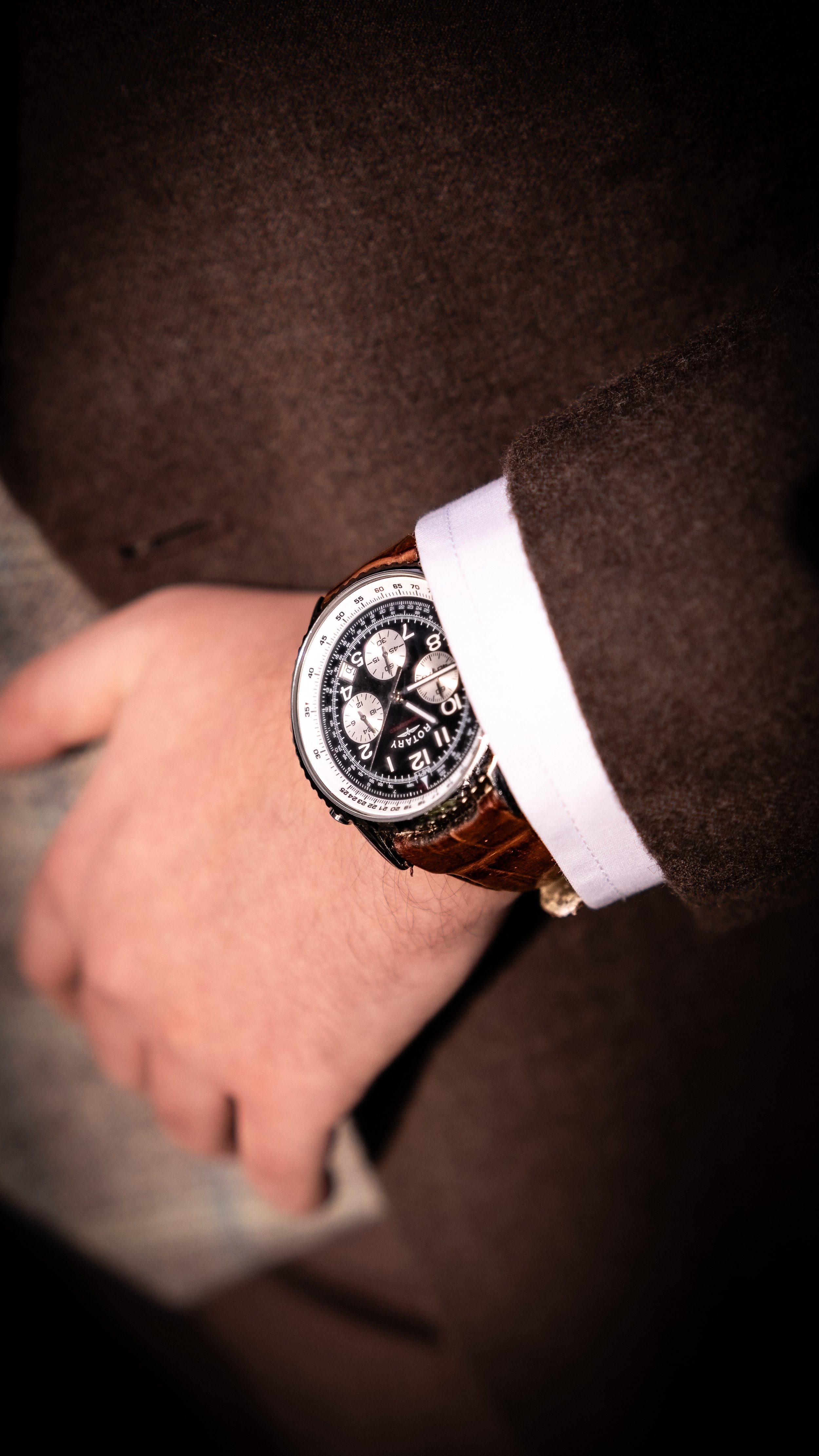 Close-up of a person's wrist with a brown leather strap watch, partially covered by a white shirt cuff. The person wears a dark brown coat.