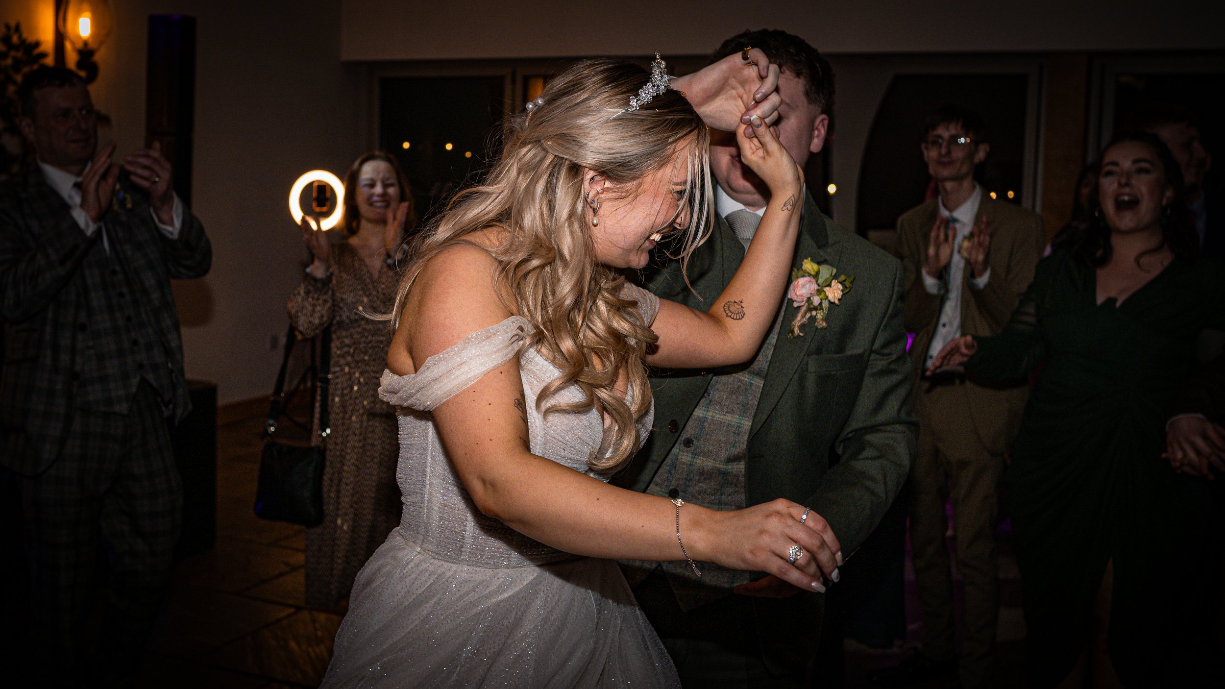A bride and groom dancing closely at their wedding reception, surrounded by happy guests clapping and smiling.