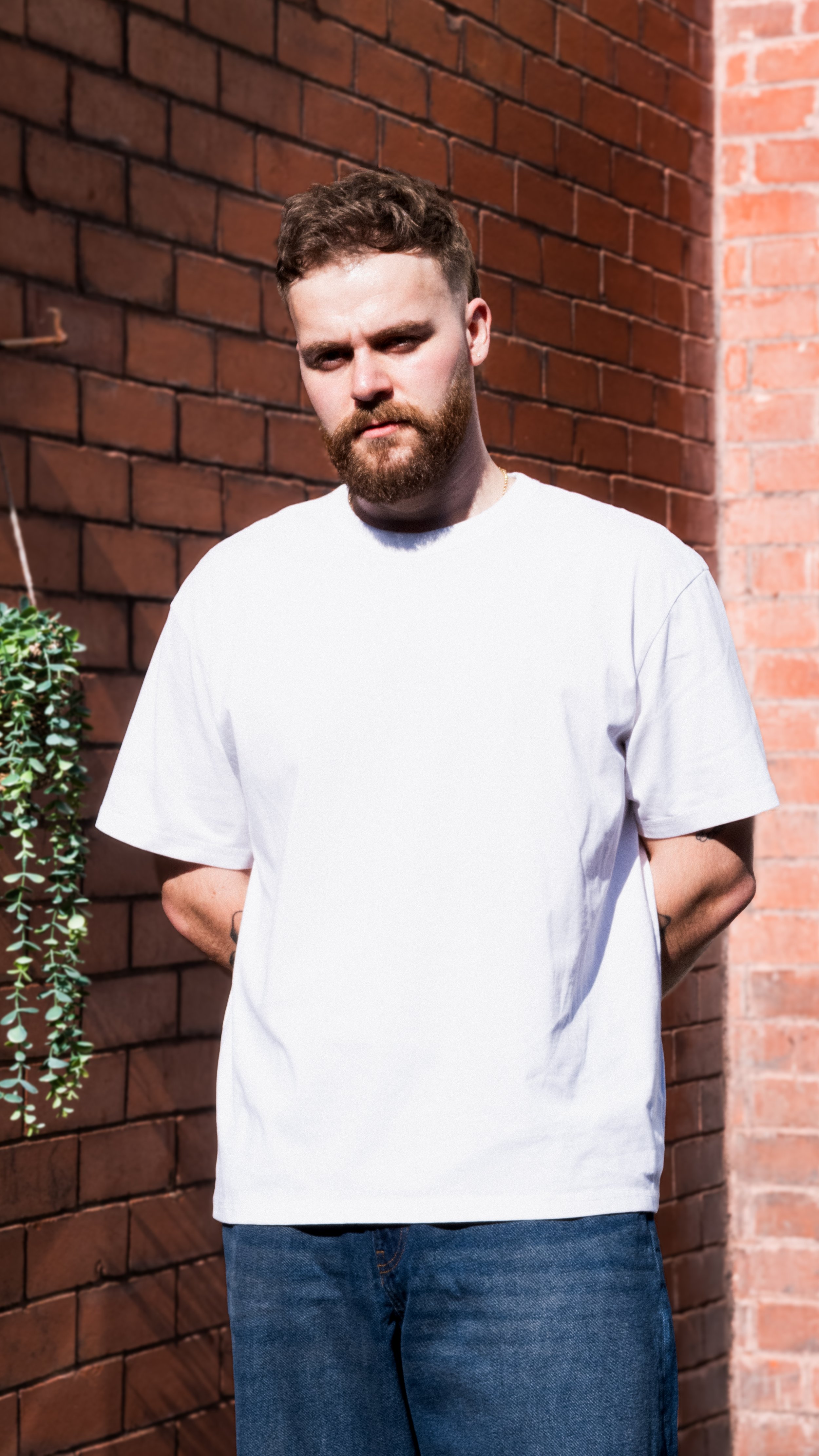 A man with short curly hair and a beard stands outdoors against a brick wall, wearing a plain white T-shirt and jeans.