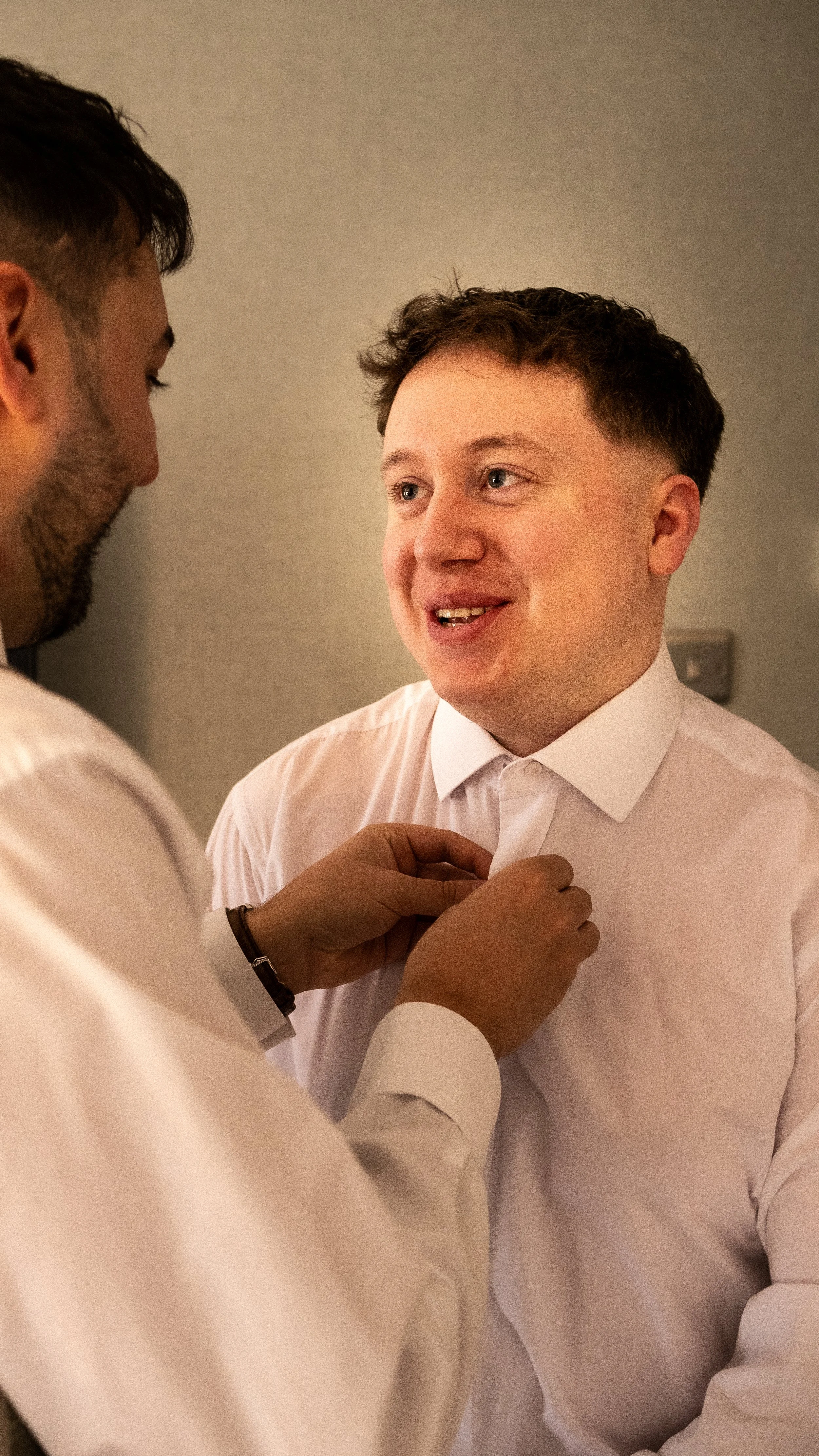 A man in a white shirt is being helped to button his collar by another person. They are indoors with a plain background.