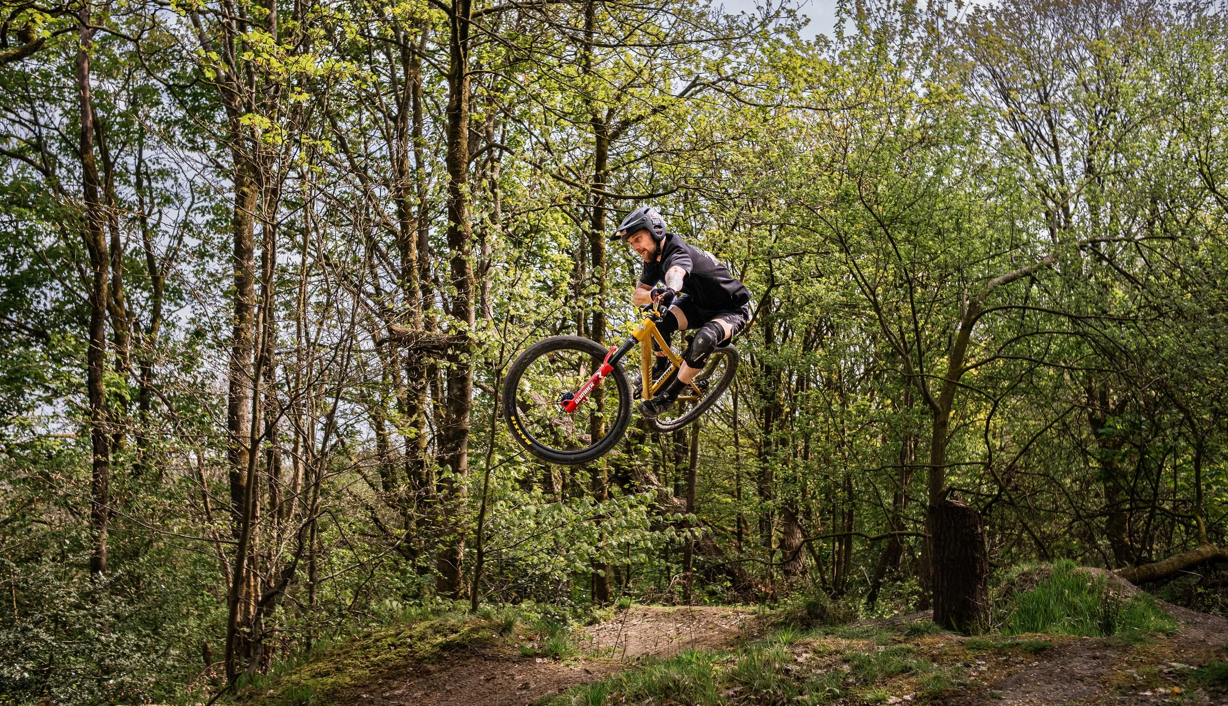 A person roller skiing with poles, airborne in the air amidst a forest with green trees and a dirt trail below.