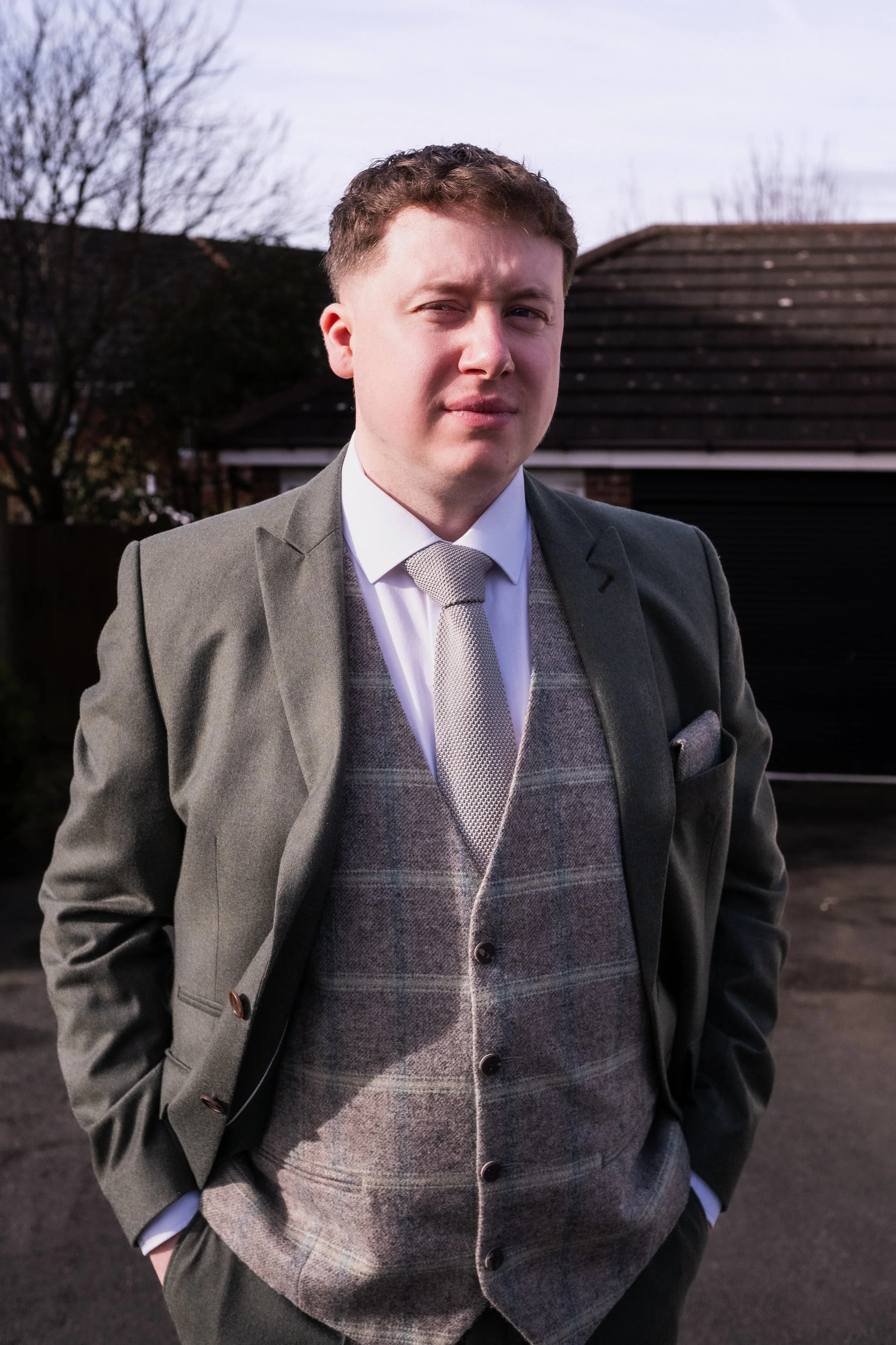 A young man in a suit standing outdoors next to a black garage door with trees and sky in the background.