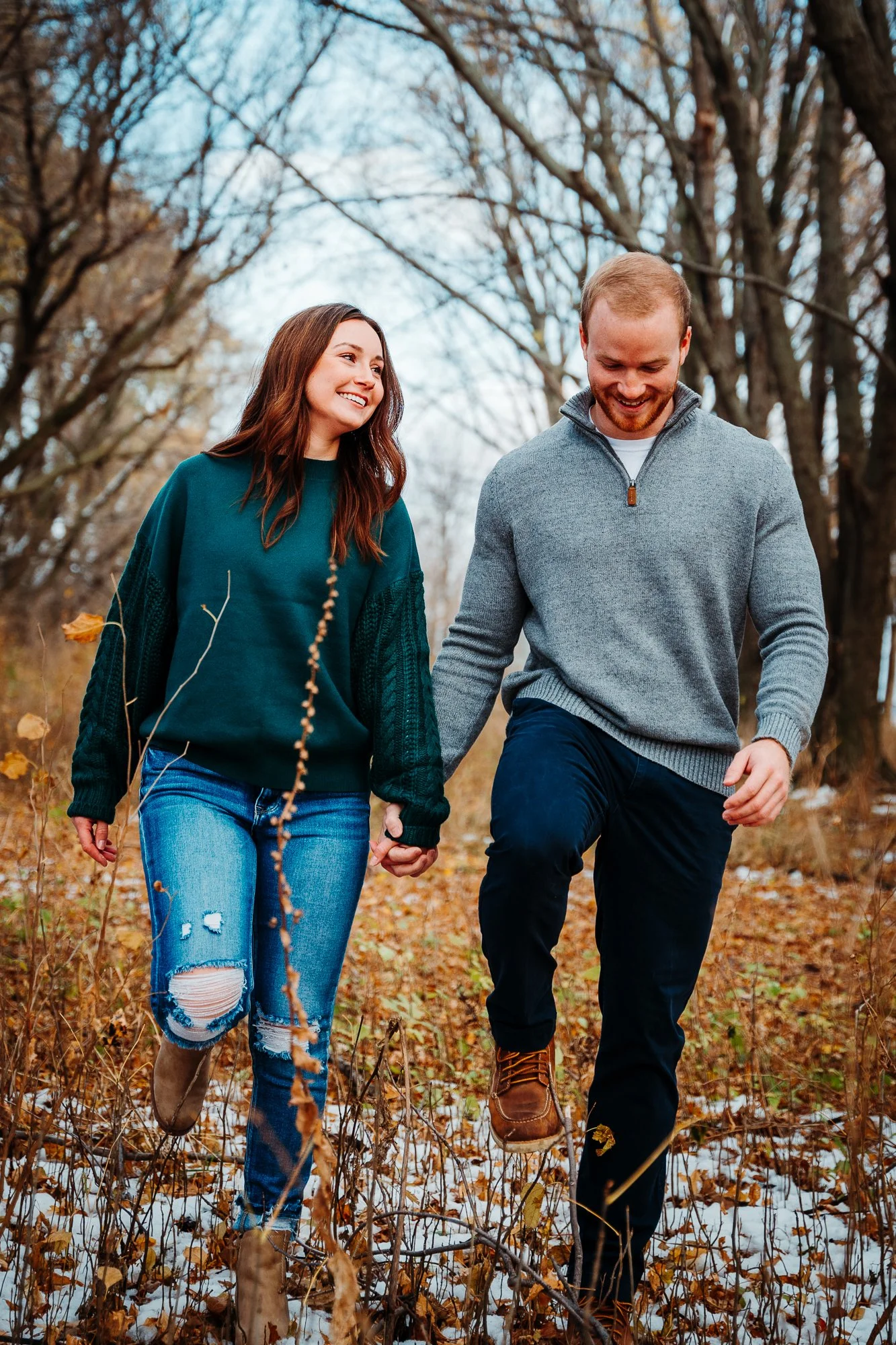 A smiling couple holding hands and walking through a wooded area on a fall day with some snow on the ground.