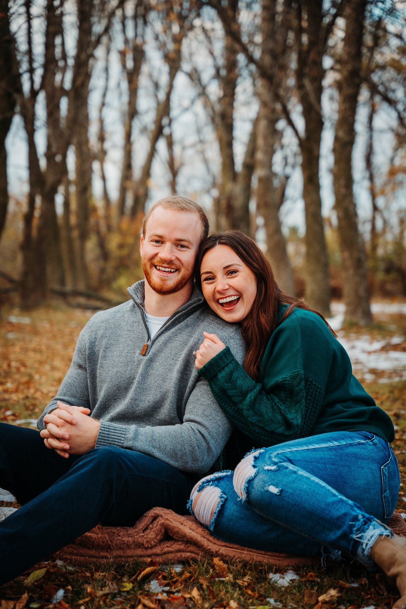 A young couple sitting on a blanket outdoors in a wooded area during fall, smiling and enjoying each other's company.