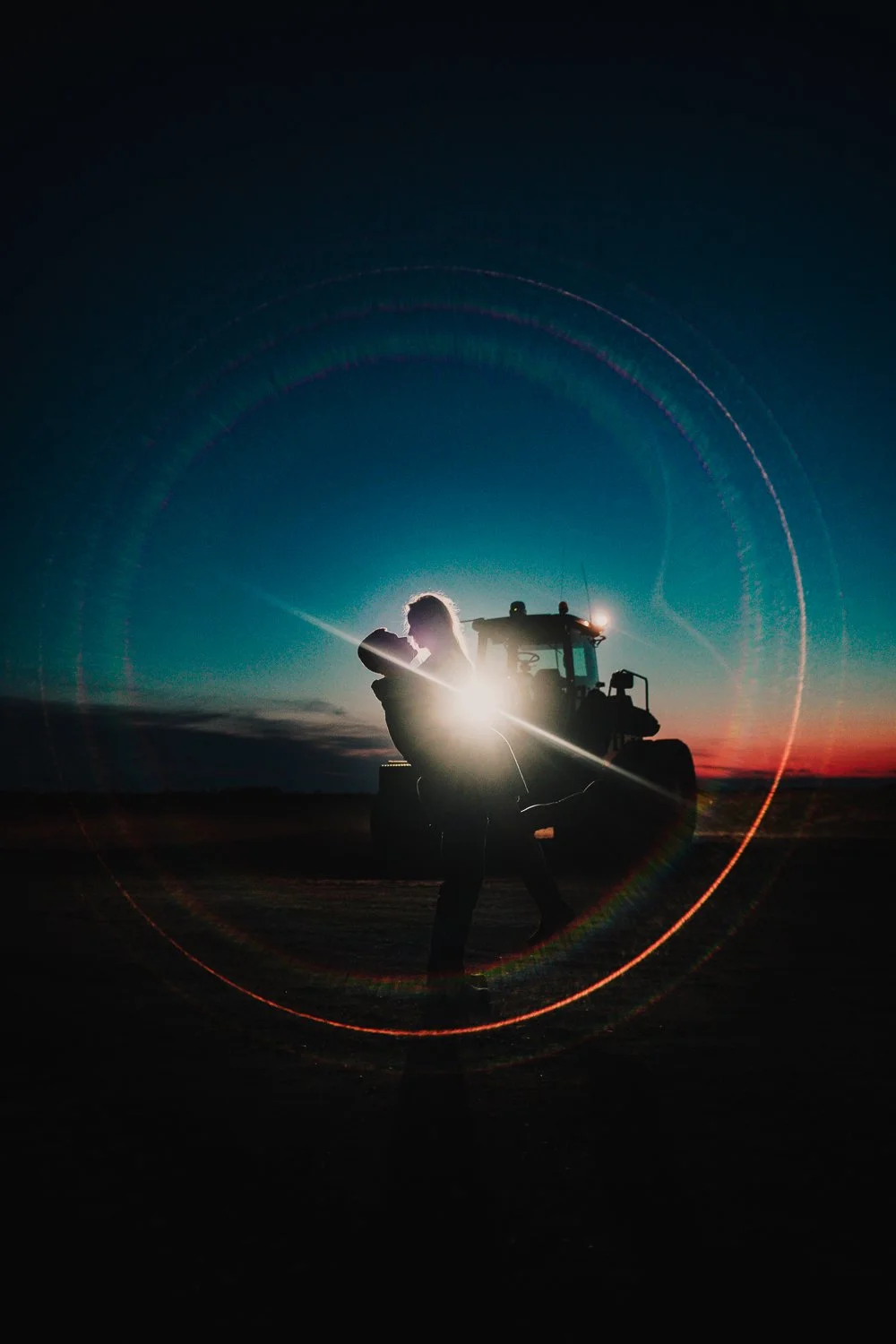 Engaged couple standing in front of a tractor during sunset with light flare and circular rainbow lens effect.