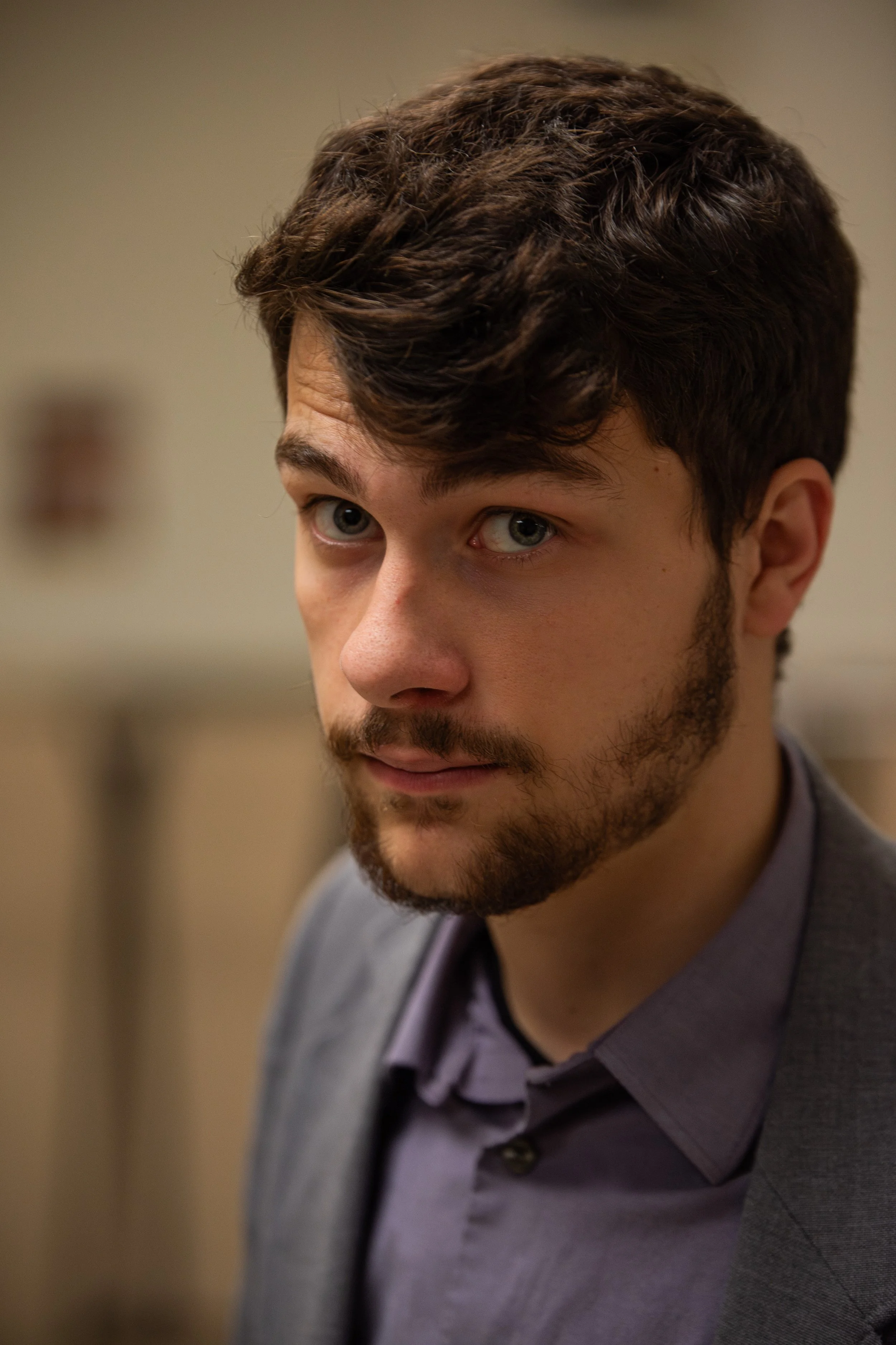 Close-up of a young man with wavy brown hair, blue eyes, and a beard, wearing a gray blazer over a purple shirt, looking to the left.