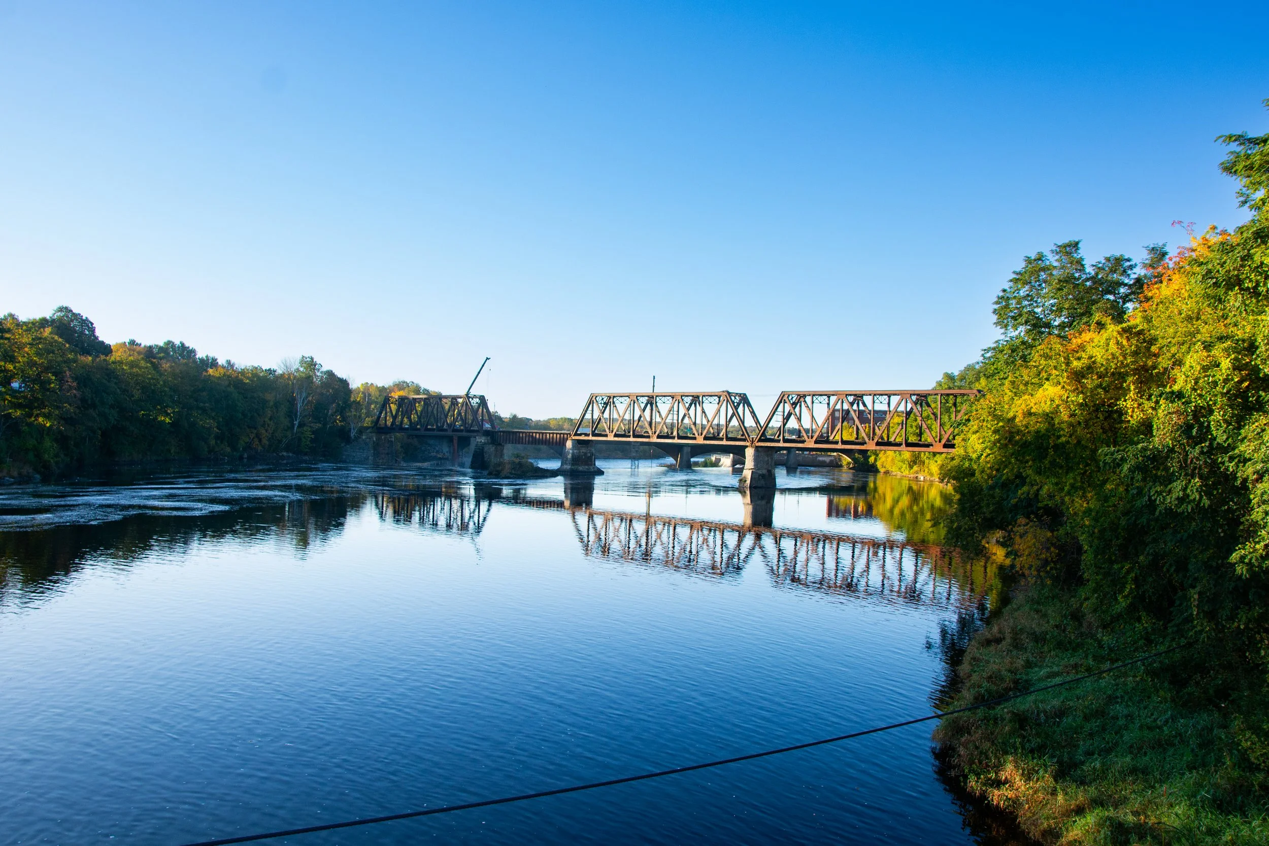 A river with a bridge crossing over it, surrounded by trees with fall foliage, and a clear blue sky above.