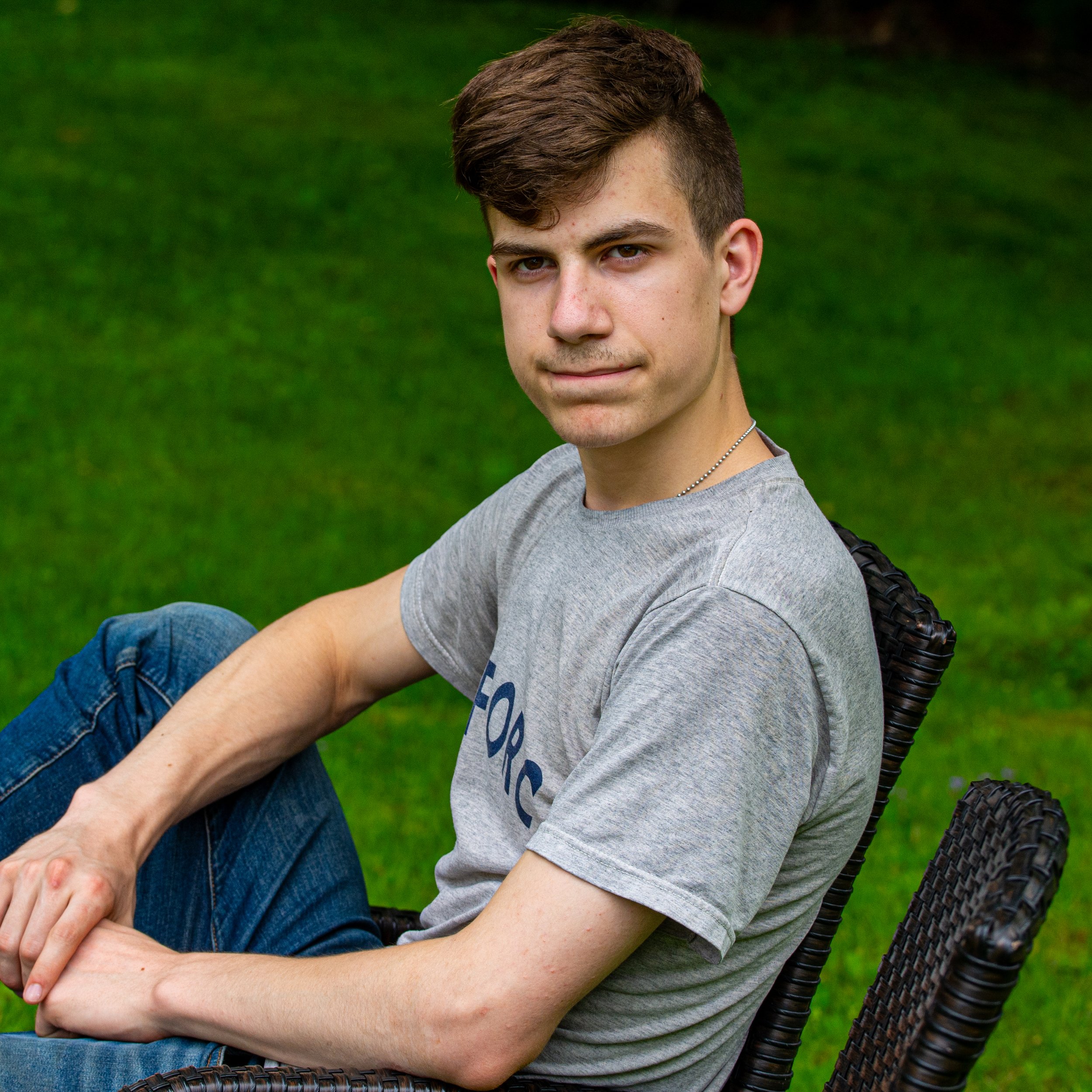 A young man with short brown hair and light skin sitting outdoors on a wicker chair with green grass in the background, wearing a gray T-shirt and blue jeans.