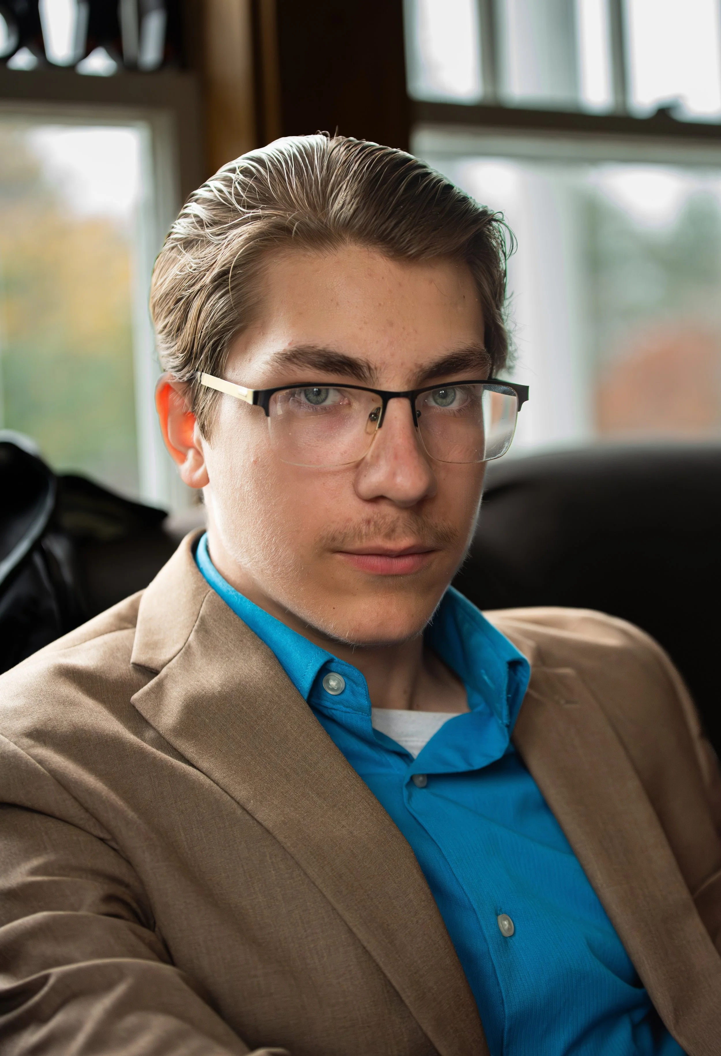 Close-up of a young man with slicked-back hair, wearing glasses, a blue dress shirt, and a tan blazer, sitting in a vehicle with large windows showing autumn trees outside.
