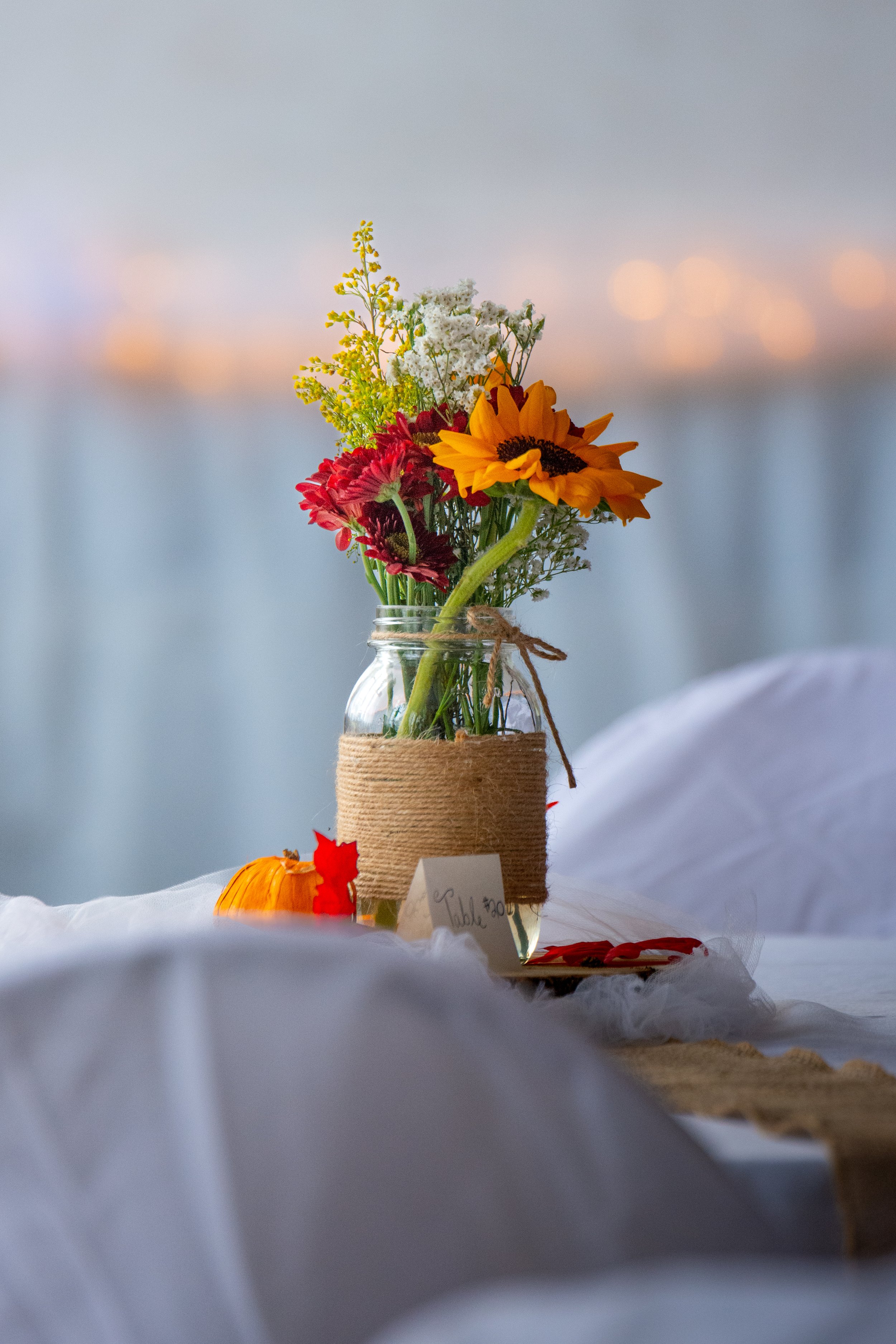 A small bouquet of flowers in a clear glass jar wrapped with twine, placed on a white fabric surface with a small pumpkin and some red leaves nearby, against a blurred background with soft lights.