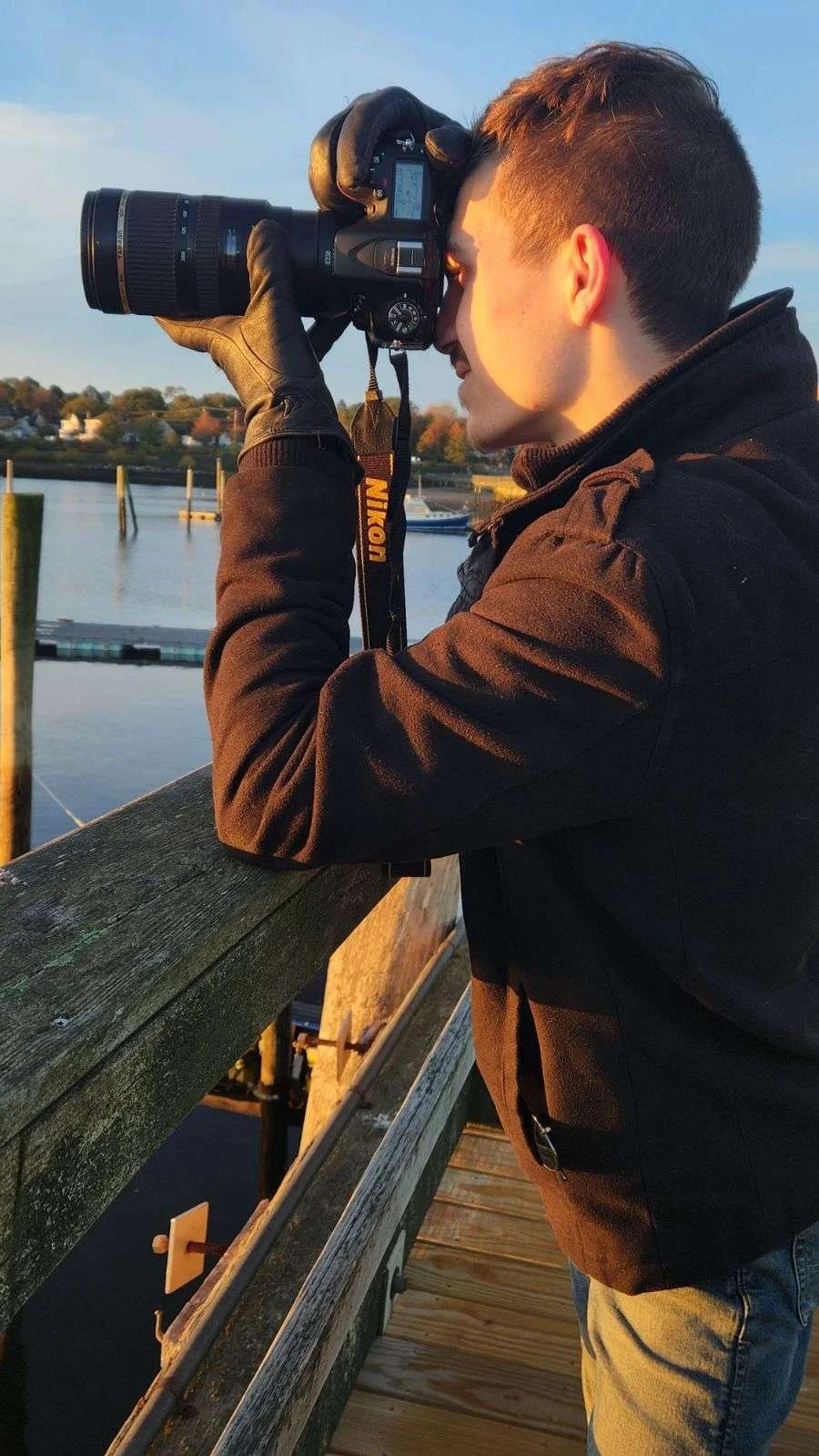A person with short brown hair wearing a black jacket and black gloves is taking a photograph with a Nikon camera on a wooden dock by the water during sunset.