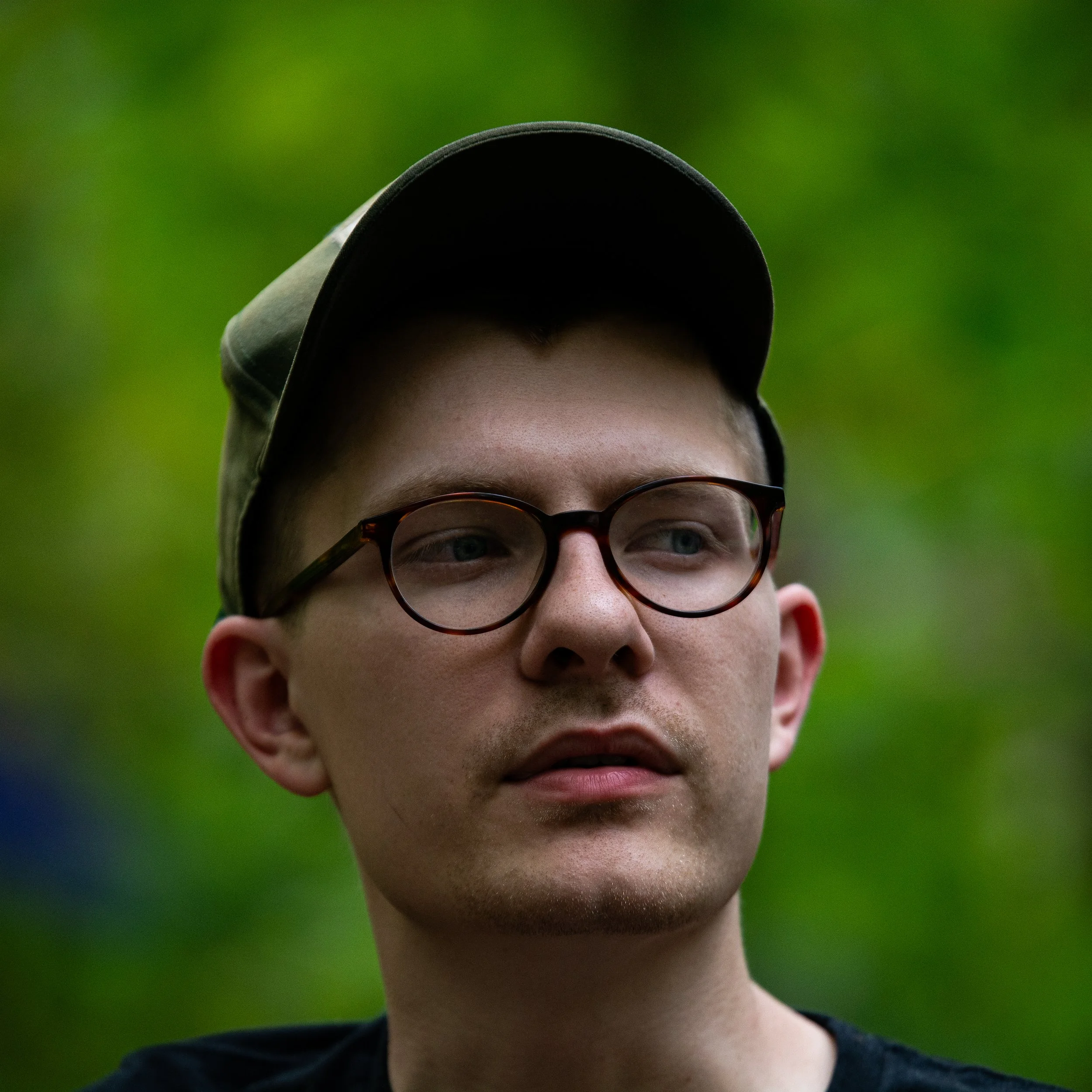 Close-up of an individual wearing glasses and a cap, with a blurred green background.