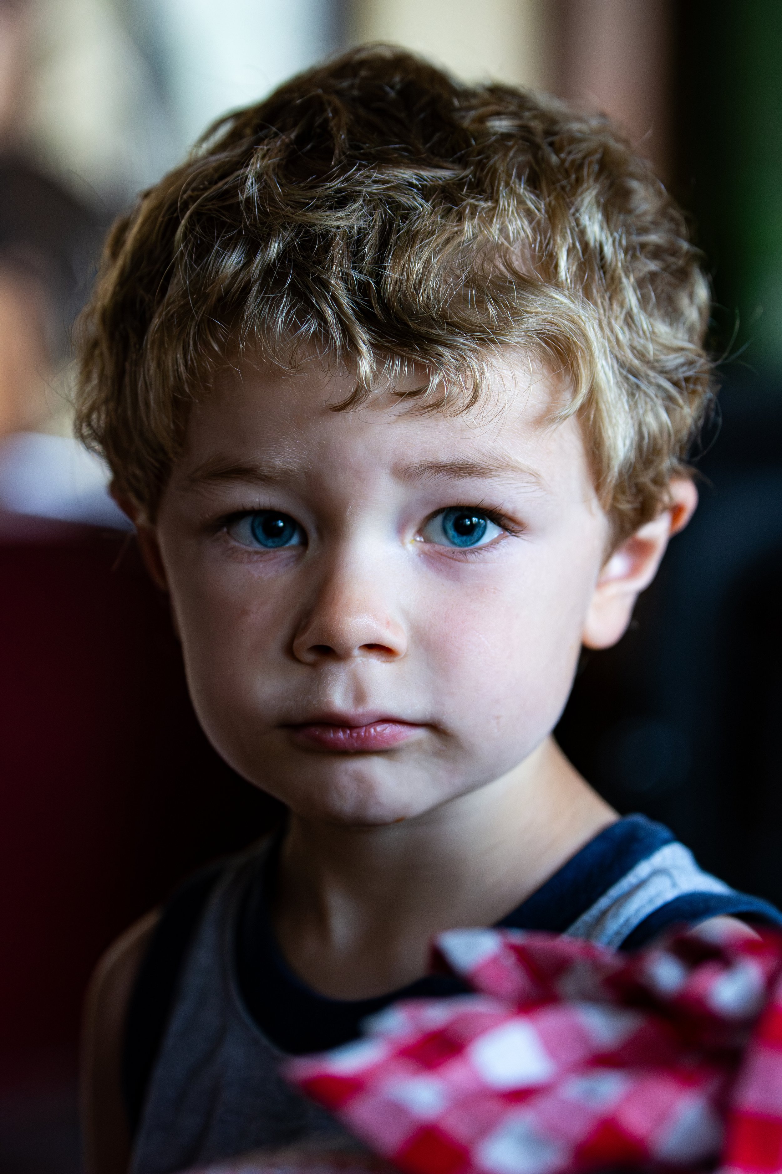 Close-up of a young child with curly hair and blue eyes looking directly at the camera. The child is wearing a striped shirt and is next to a gift wrapped in red and white checkered paper with a pink ribbon.