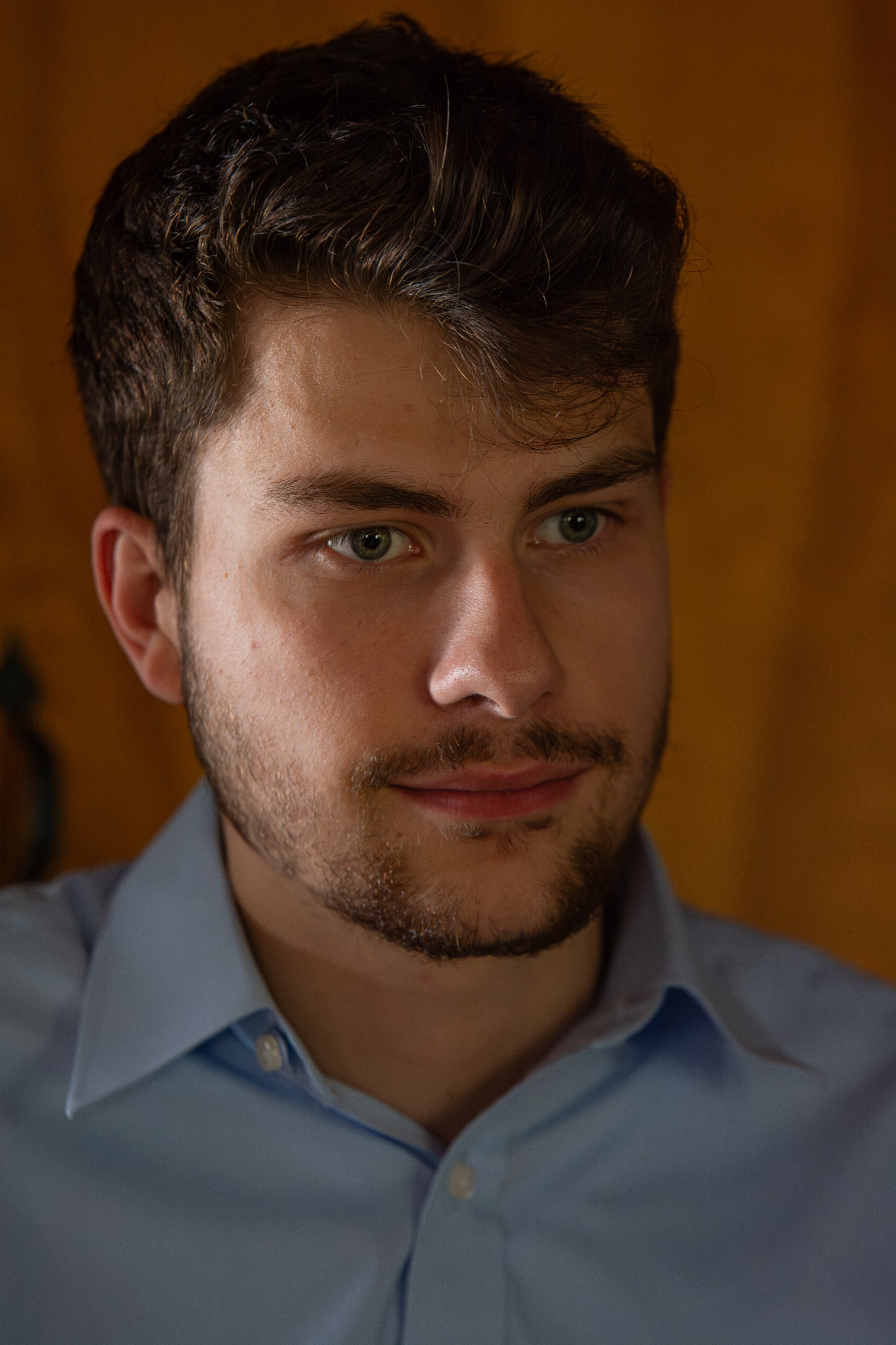 Close-up of a young man with dark hair, green eyes, and light beard, wearing a light blue shirt, indoors with warm background lighting.